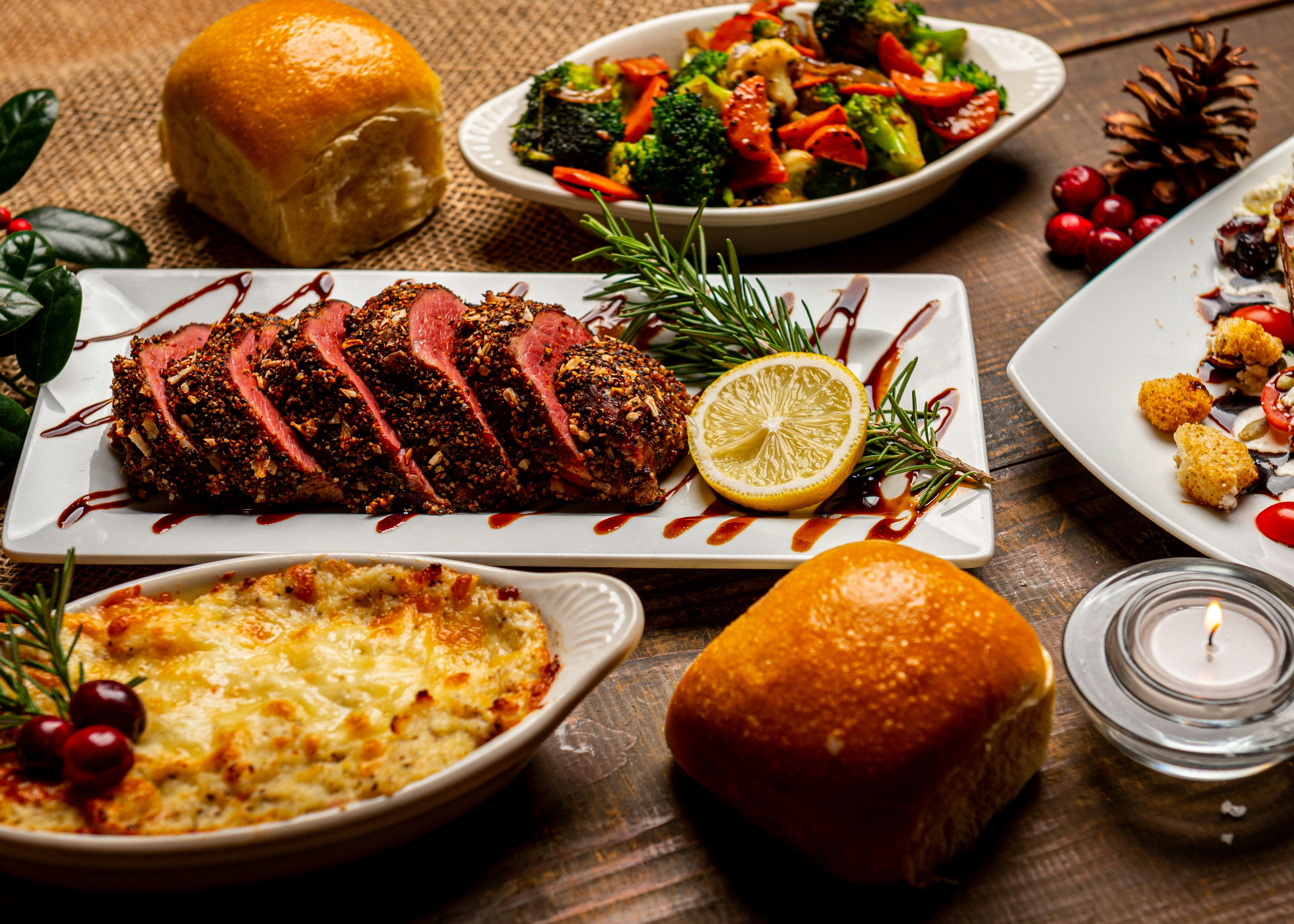 A festive dinner spread featuring sliced roast beef, bread rolls, baked casserole, roasted vegetables, and garnishes.