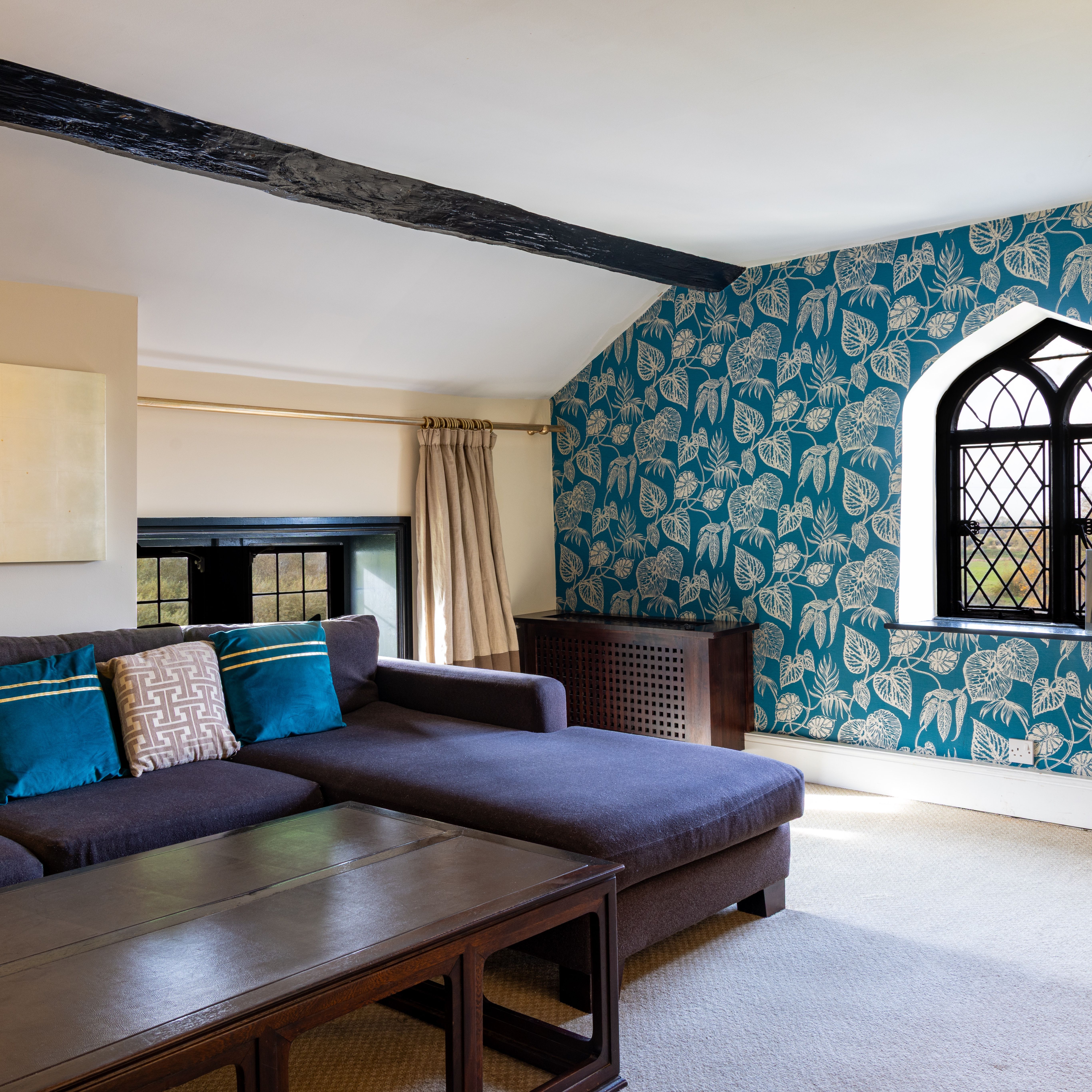 A cozy living room with a sectional sofa, blue and patterned cushions, wooden coffee table, and a wall with blue floral wallpaper and an arched window.