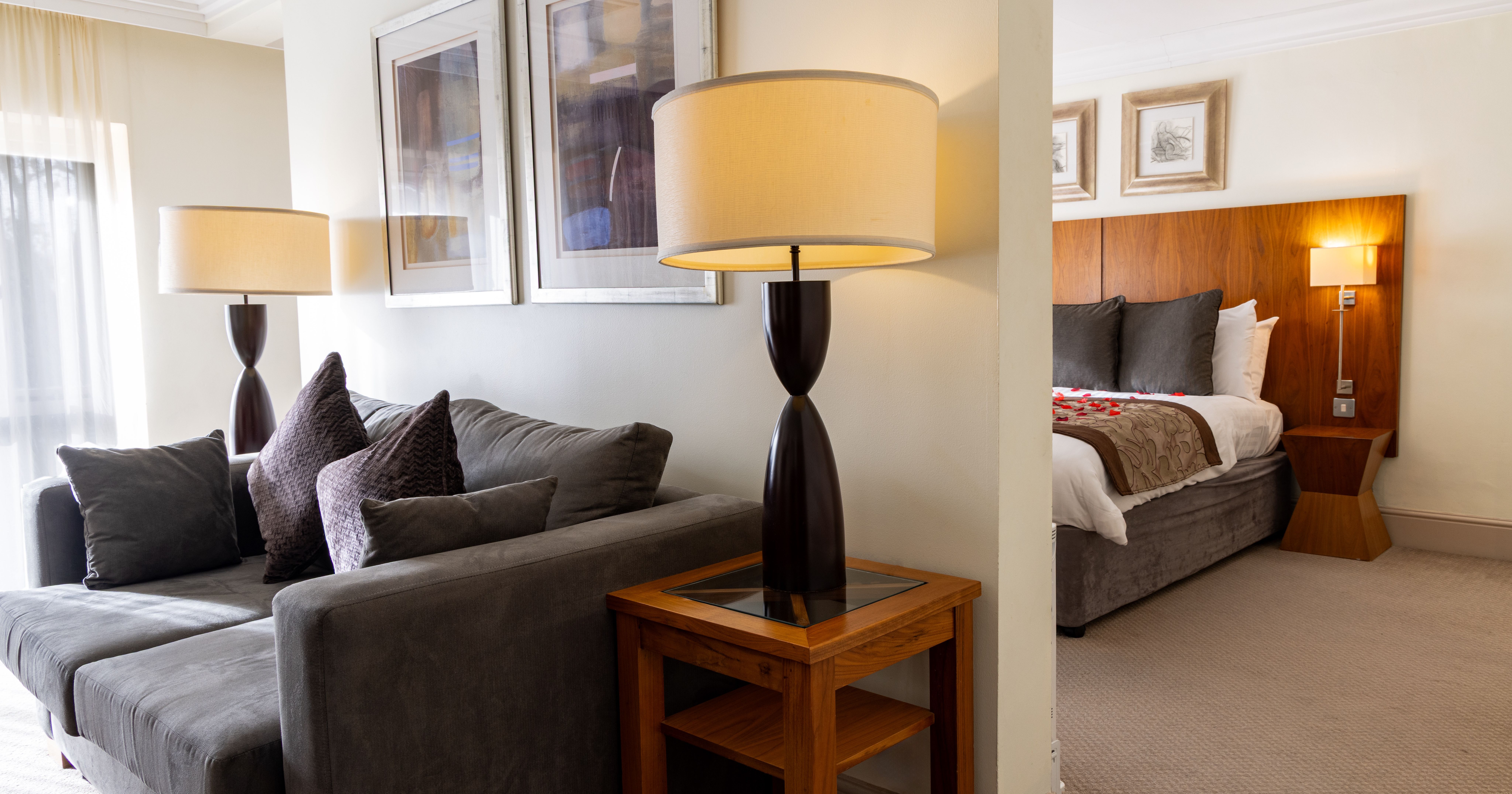 Modern hotel suite with living area, grey sofa, table lamp, and bedroom with bed in the background