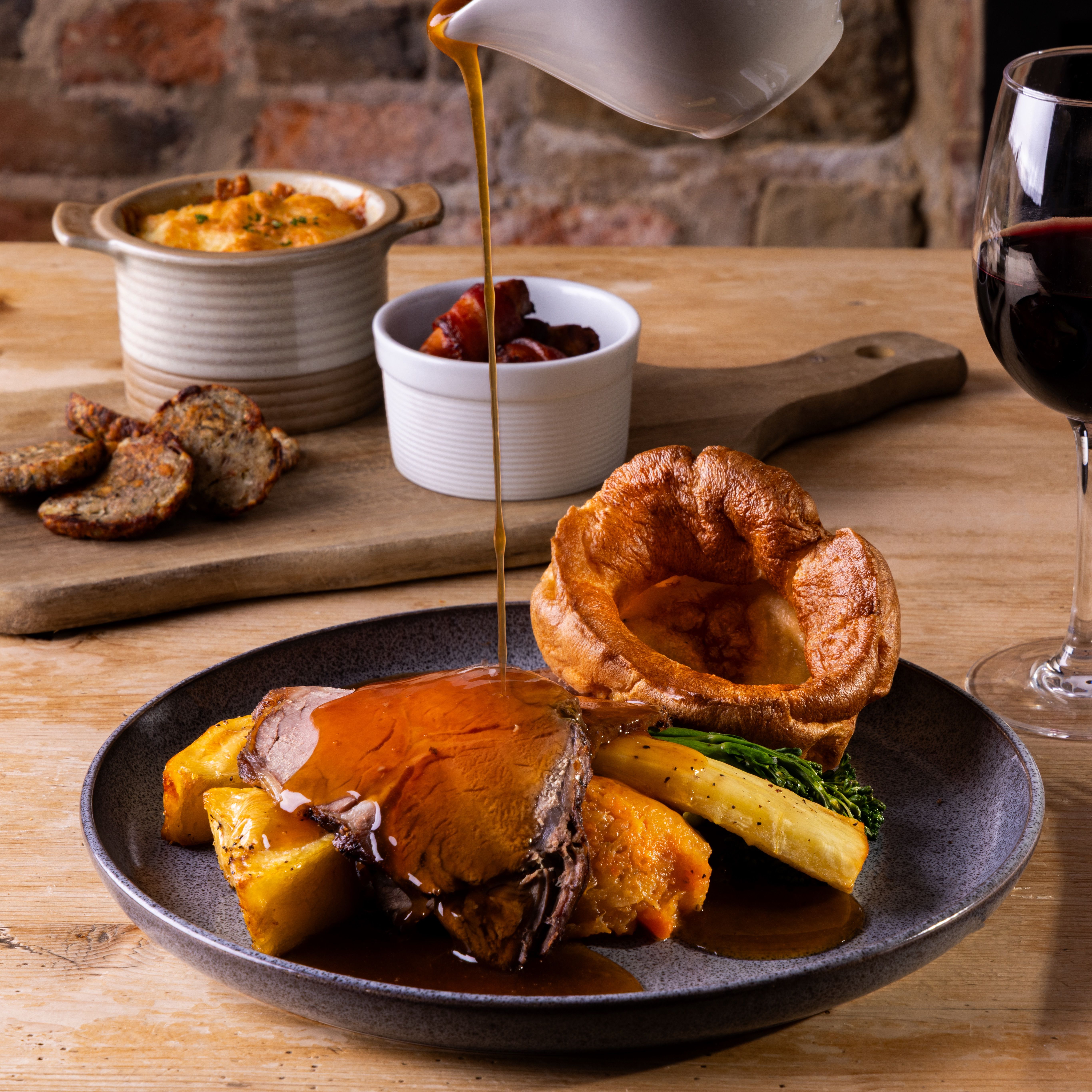 Sunday roast dinner with Yorkshire pudding, roast beef, vegetables, and gravy being poured, accompanied by sides and a glass of red wine.