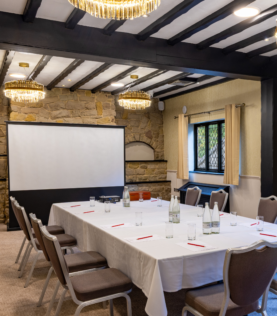 A well-lit meeting room with a large table set for a conference, surrounded by chairs, a presentation screen, and decorative stone walls.