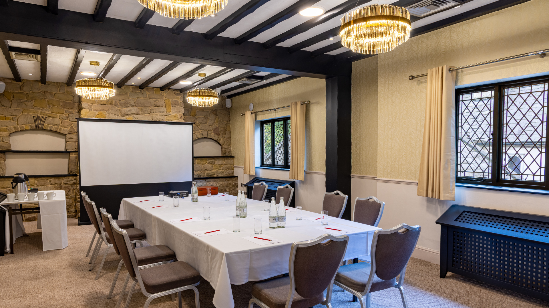 A well-lit meeting room with a large table set for a conference, surrounded by chairs, a presentation screen, and decorative stone walls.