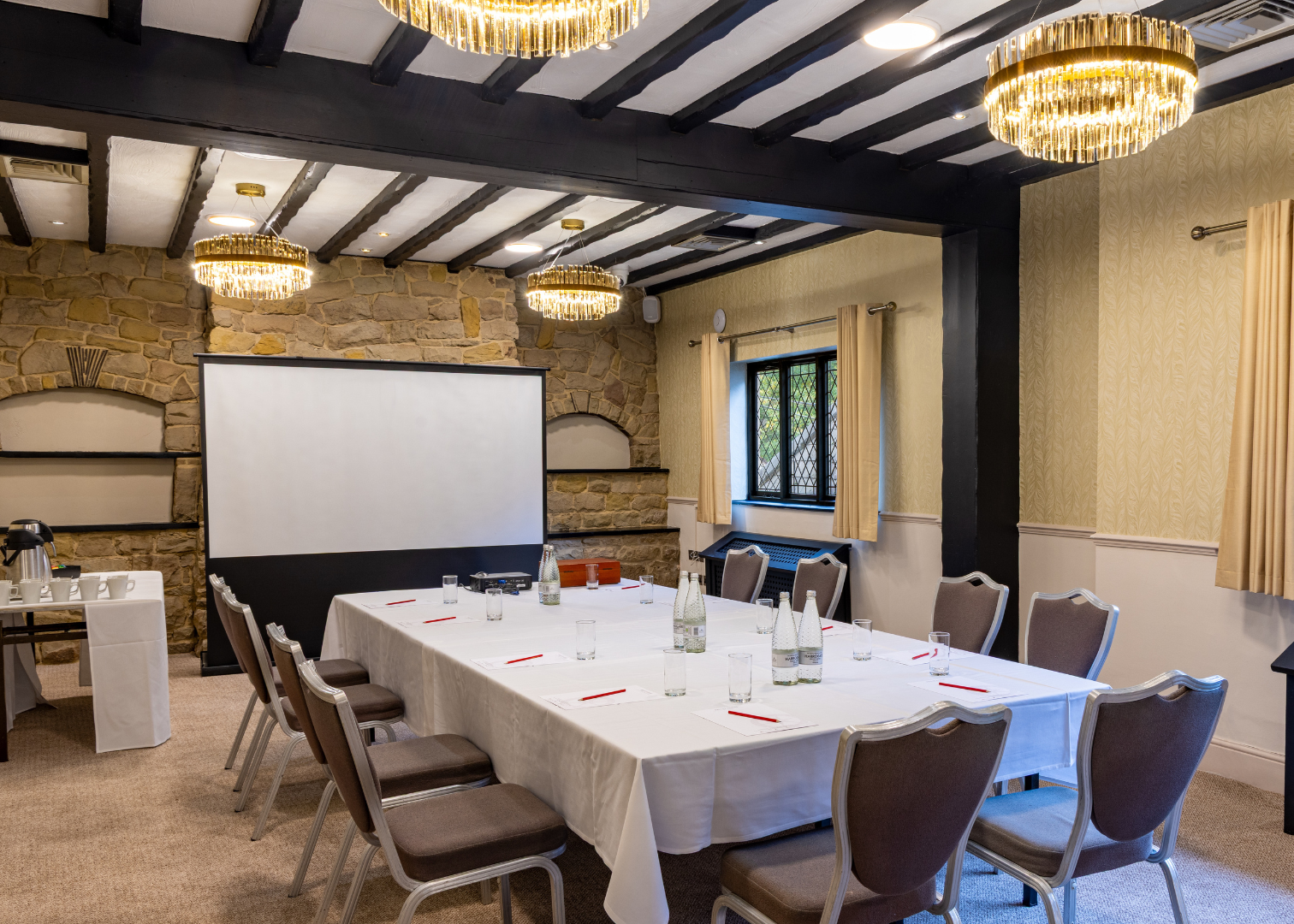 A well-lit meeting room with a large table set for a conference, surrounded by chairs, a presentation screen, and decorative stone walls.