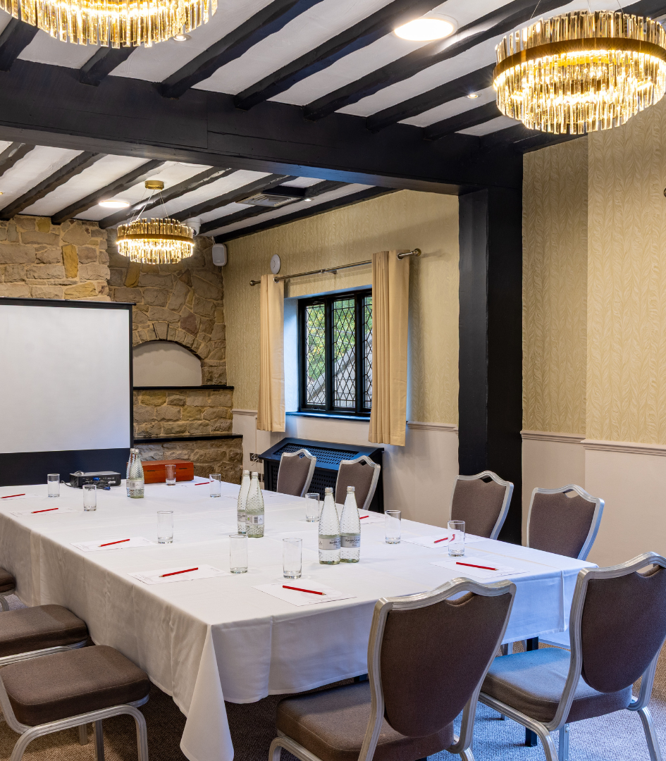 A well-lit meeting room with a large table set for a conference, surrounded by chairs, a presentation screen, and decorative stone walls.
