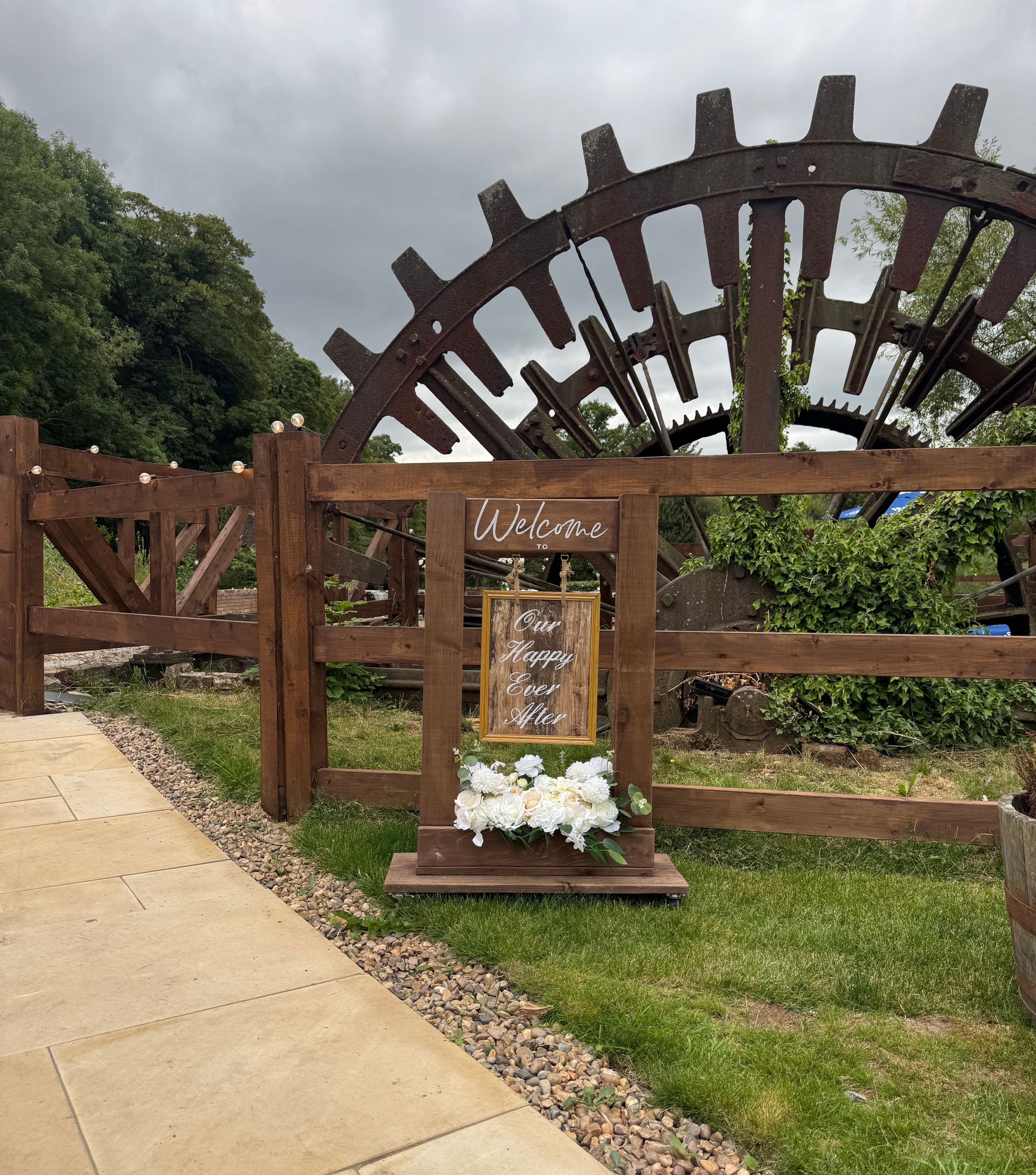 Welcome sign with flowers in front of a large water wheel and wooden fence at Kings Mills Deck.