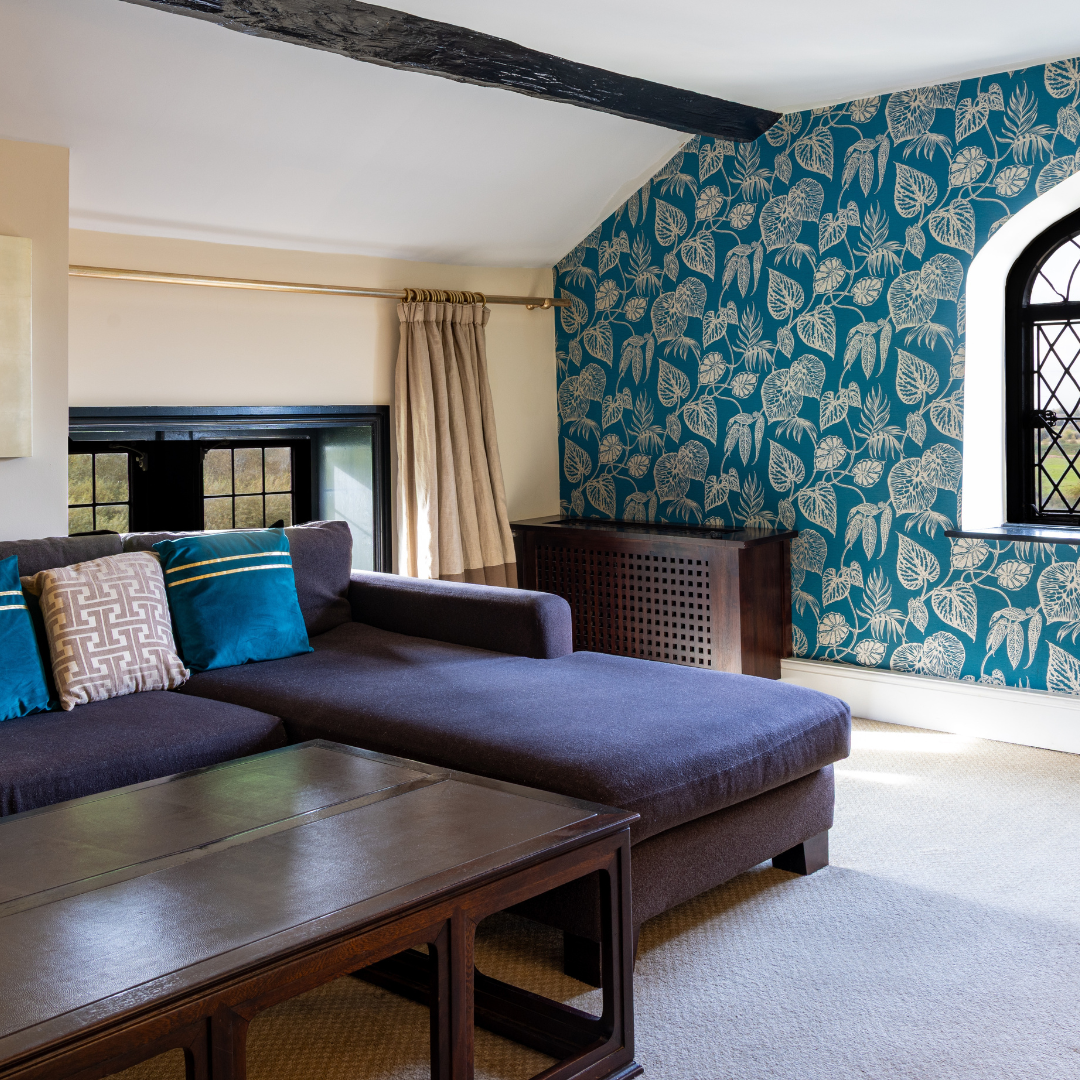 Cozy living room with a large sectional sofa, patterned pillows, wooden coffee table, and a feature wall with blue leaf-patterned wallpaper and an arched window.