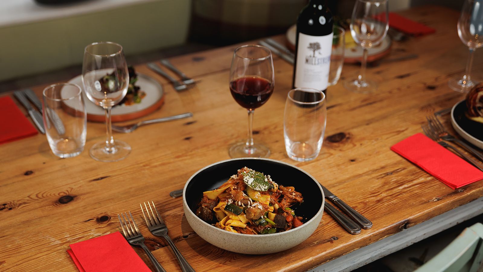 Rustic restaurant table set with a bowl of pasta, wine glasses, red napkins, and a bottle of red wine.