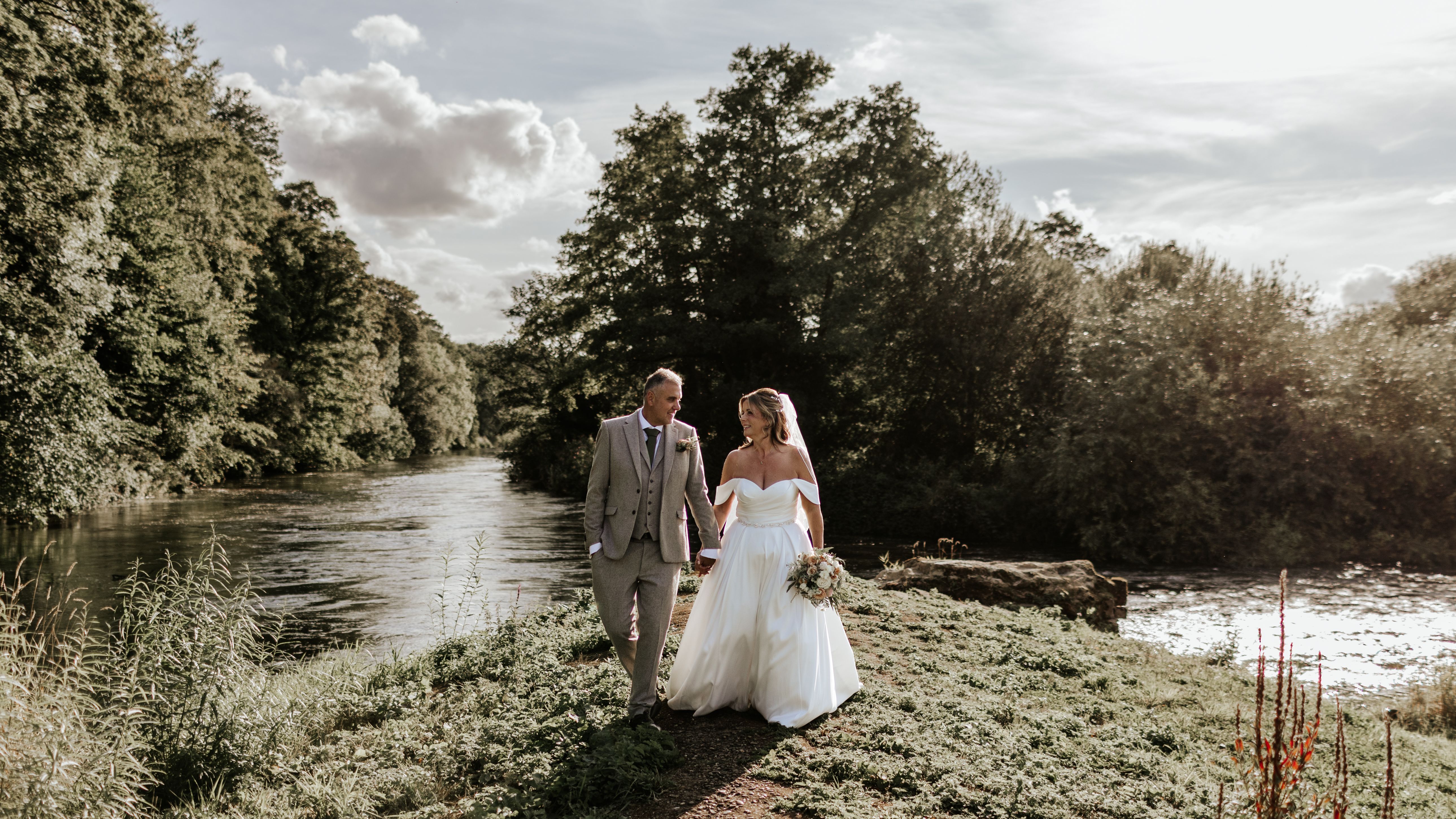 Bride and groom walking hand-in-hand alongside a river surrounded by greenery.