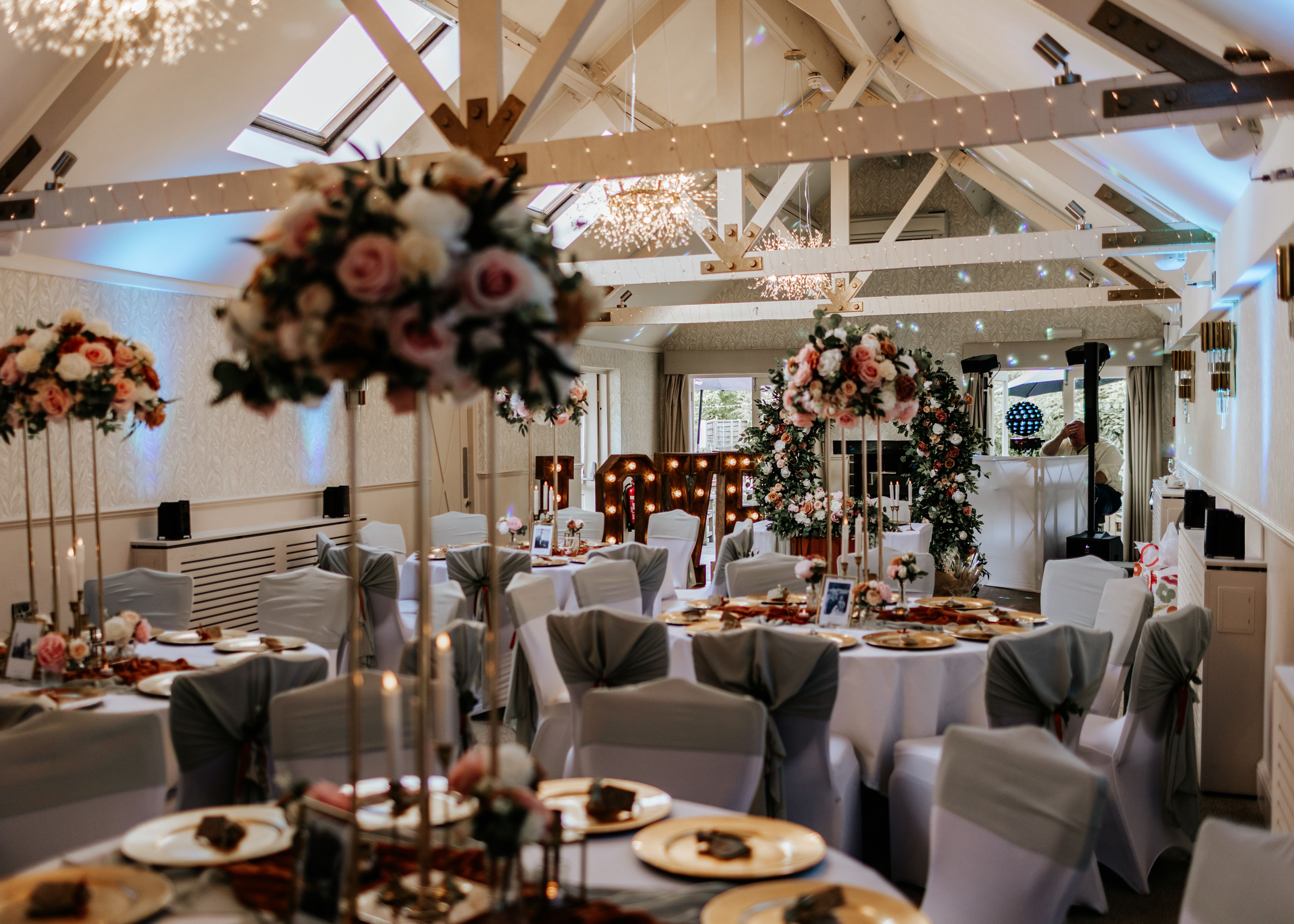 Elegant wedding reception hall decorated with floral centerpieces, round tables, white chair covers, and illuminated 'LOVE' letters.