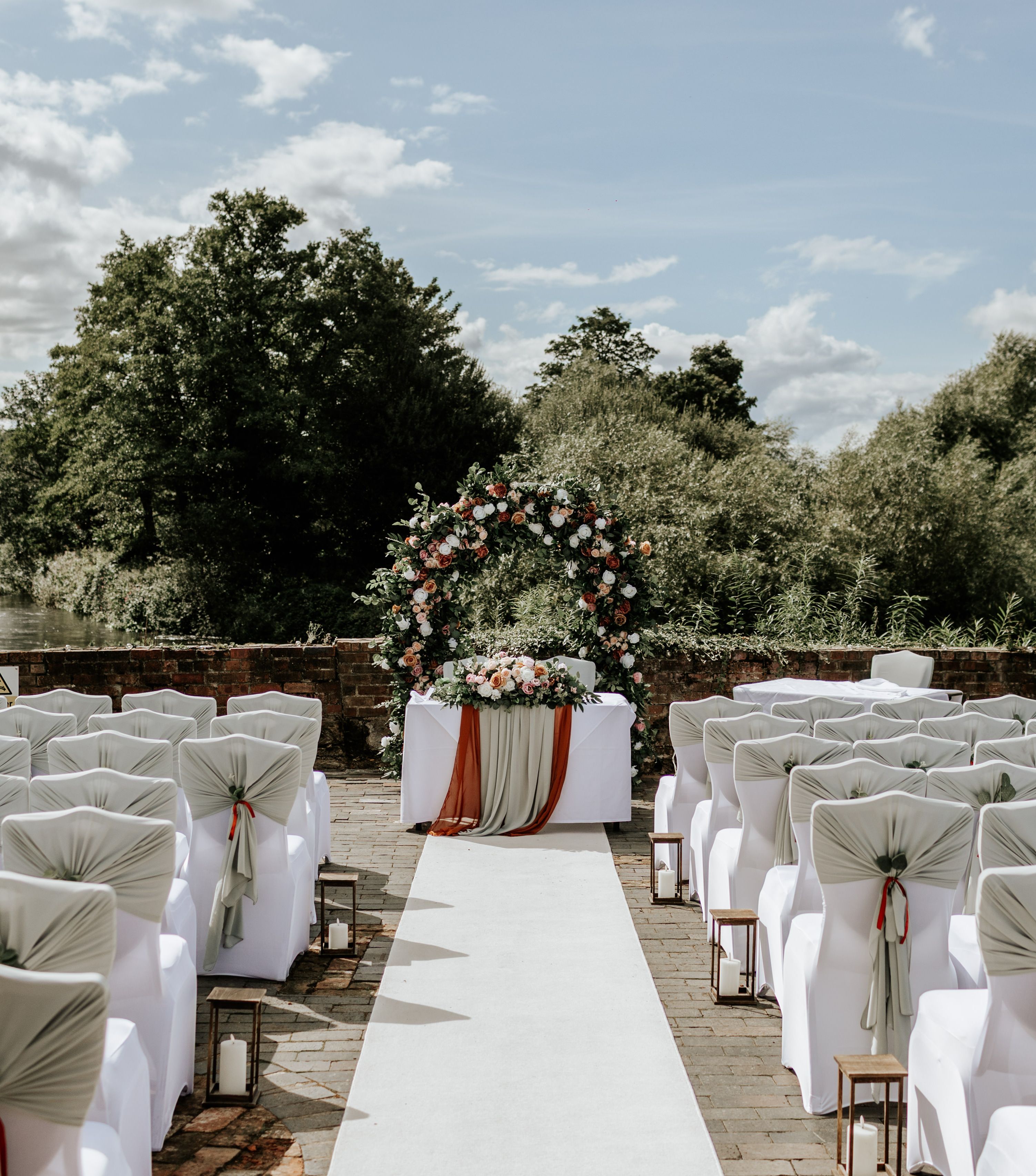 Outdoor wedding ceremony setup with decorated chairs, floral arch, and table along a river backdrop.