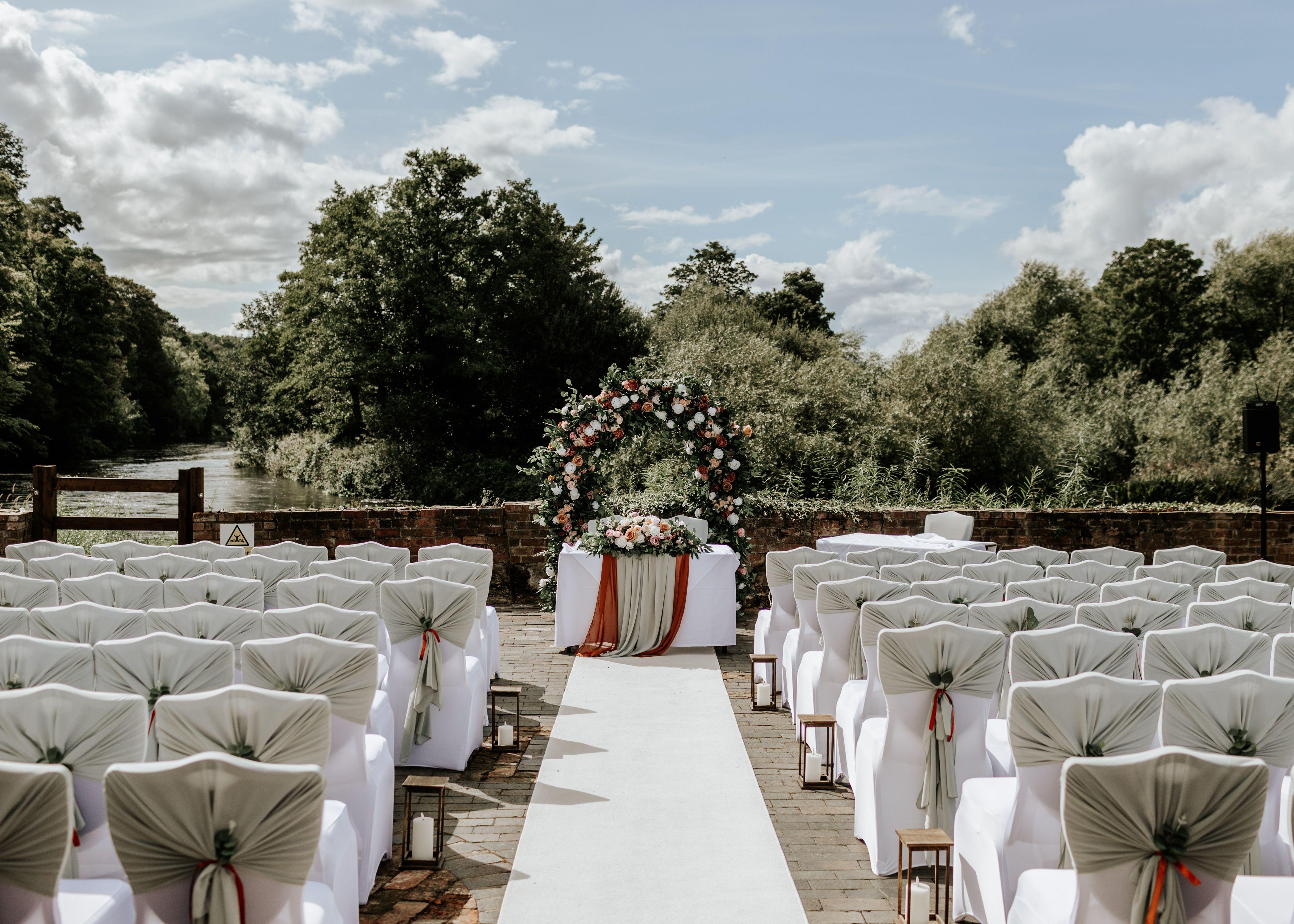Outdoor wedding ceremony setup with decorated chairs, floral arch, and table along a river backdrop.