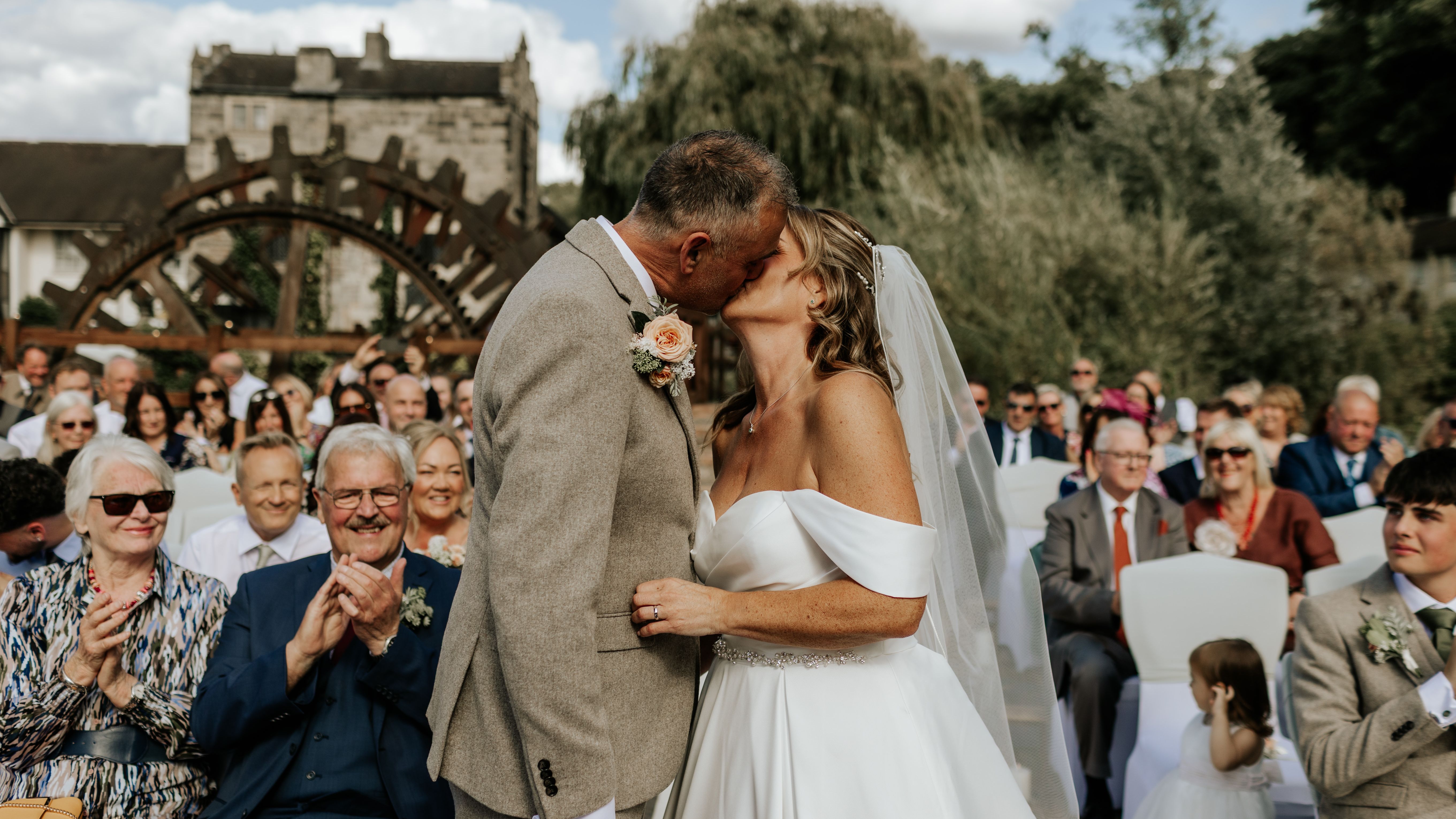 Bride and groom sharing a kiss at their outdoor wedding ceremony with guests applauding in the background.