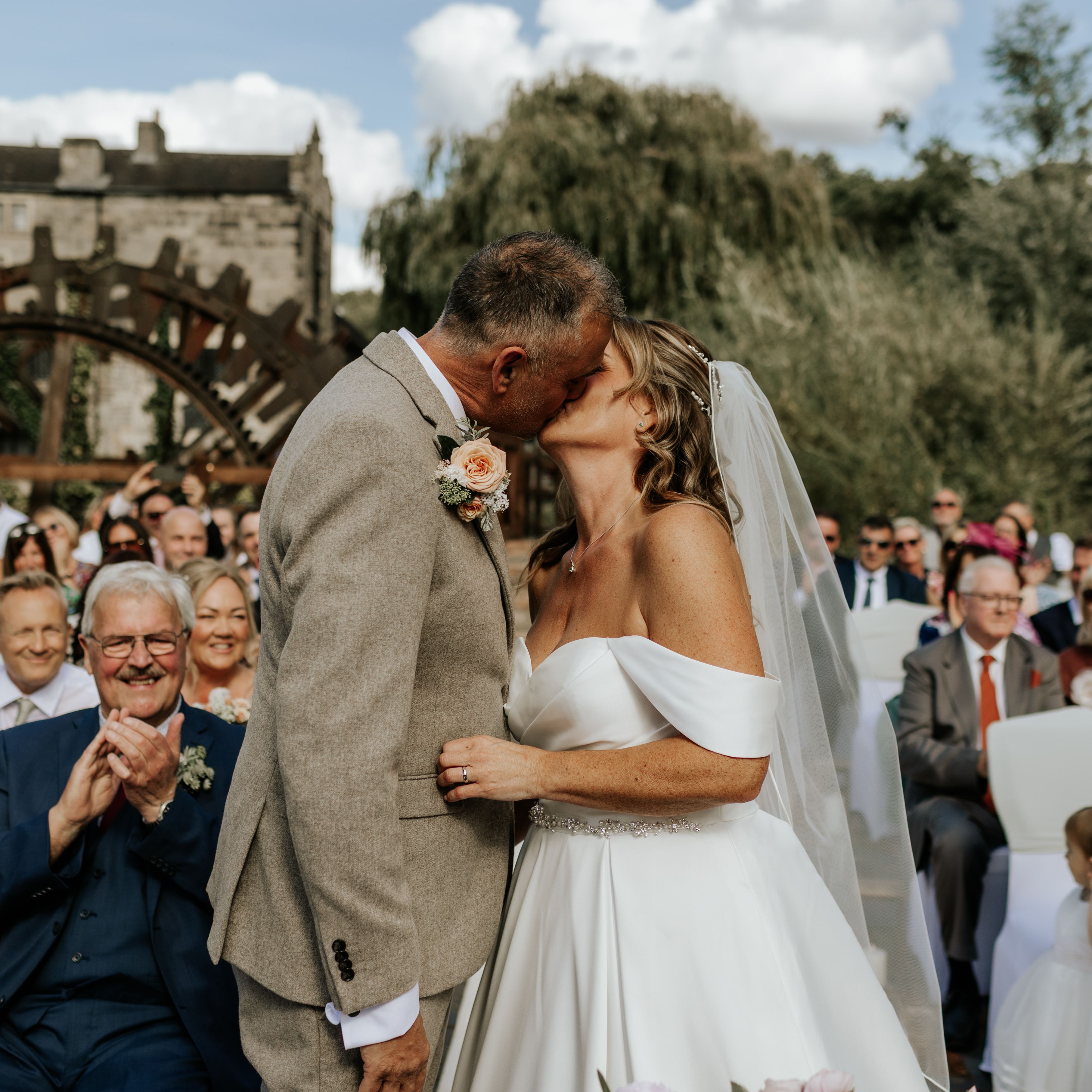 Bride and groom sharing a kiss at their outdoor wedding ceremony with guests applauding in the background.