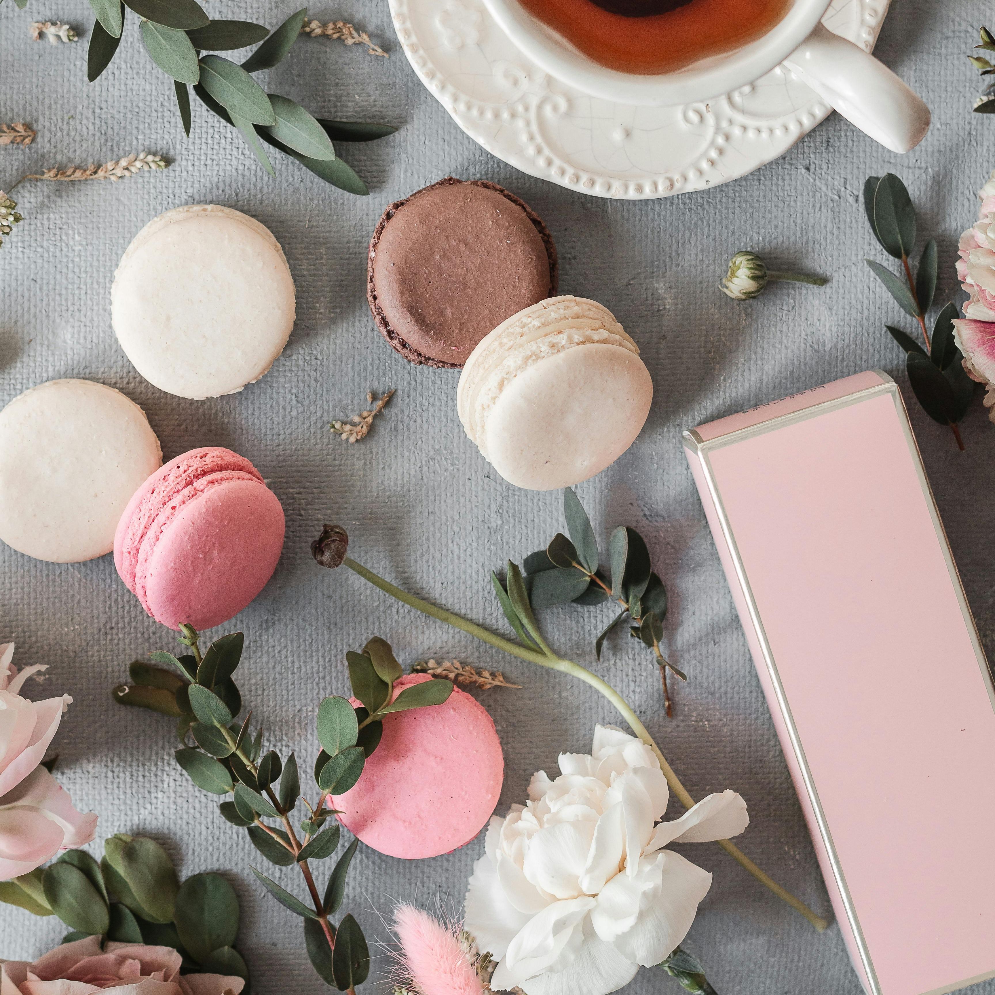 A cup of tea surrounded by assorted macarons and flowers on a textured surface.