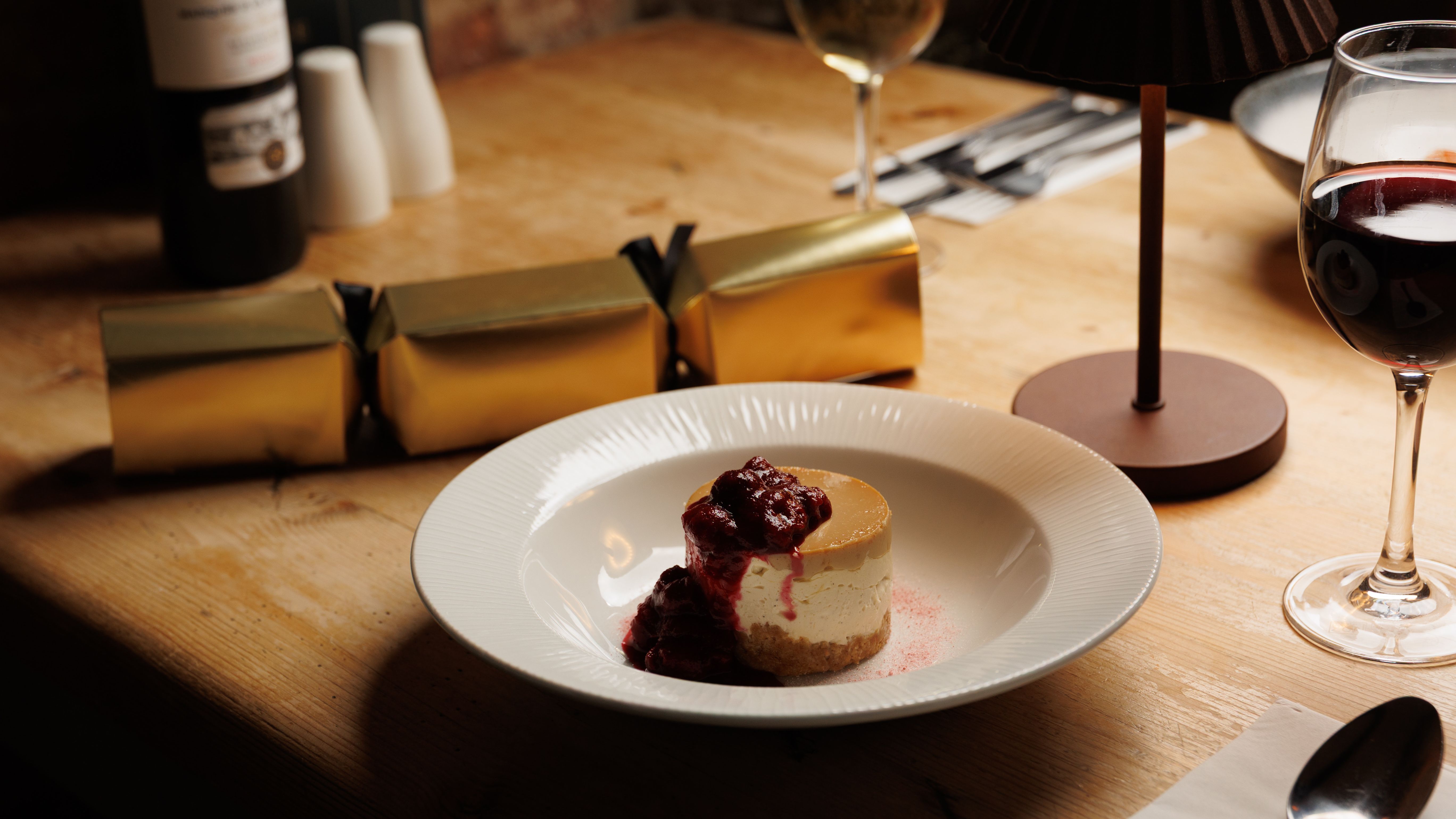 A dessert topped with berry compote on a white plate, on a wooden dining table set with wine glasses, cutlery, and gold crackers.