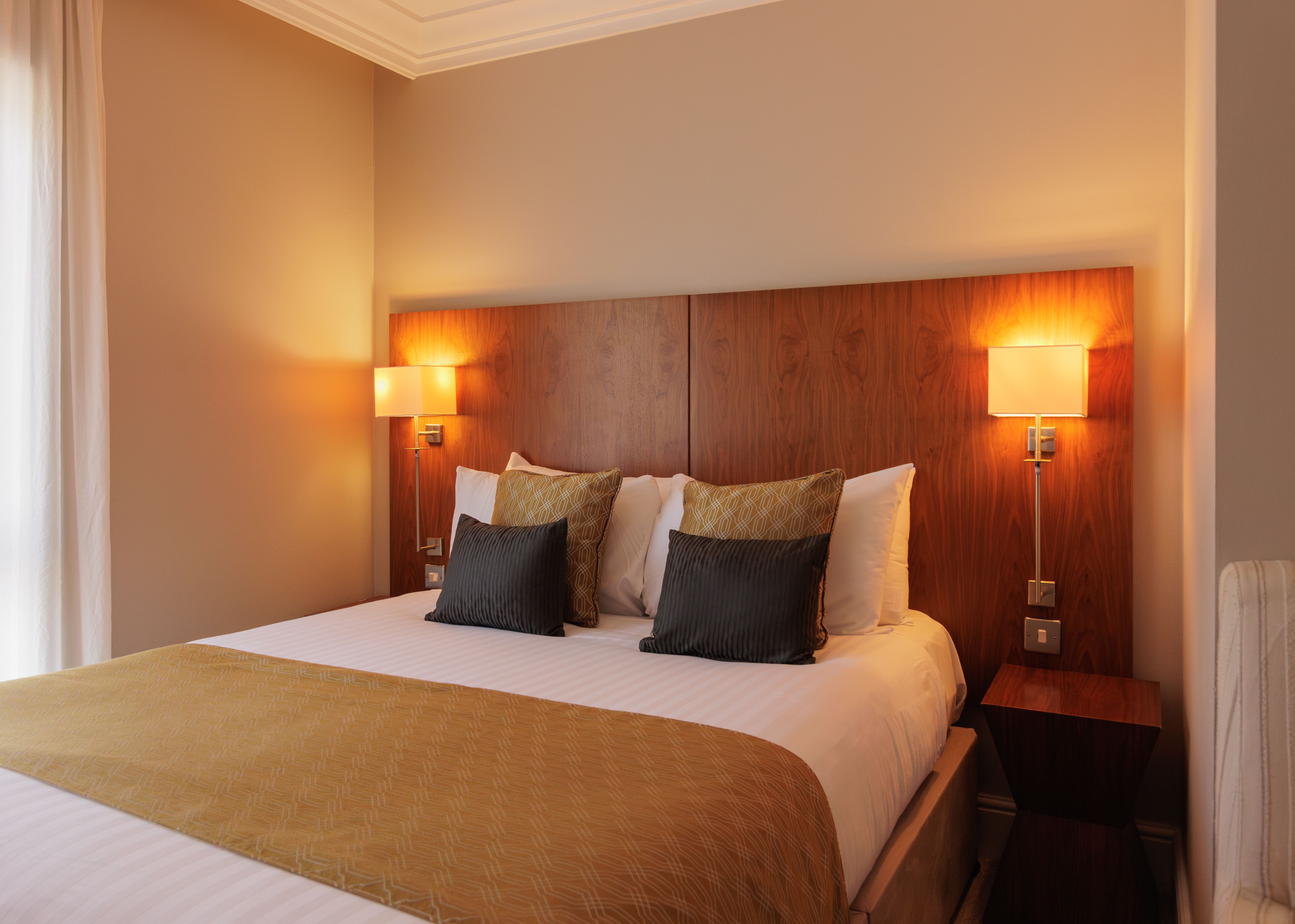 A cozy, modern hotel bedroom with a double bed, wooden headboard, bedside lamps, and decorative pillows.