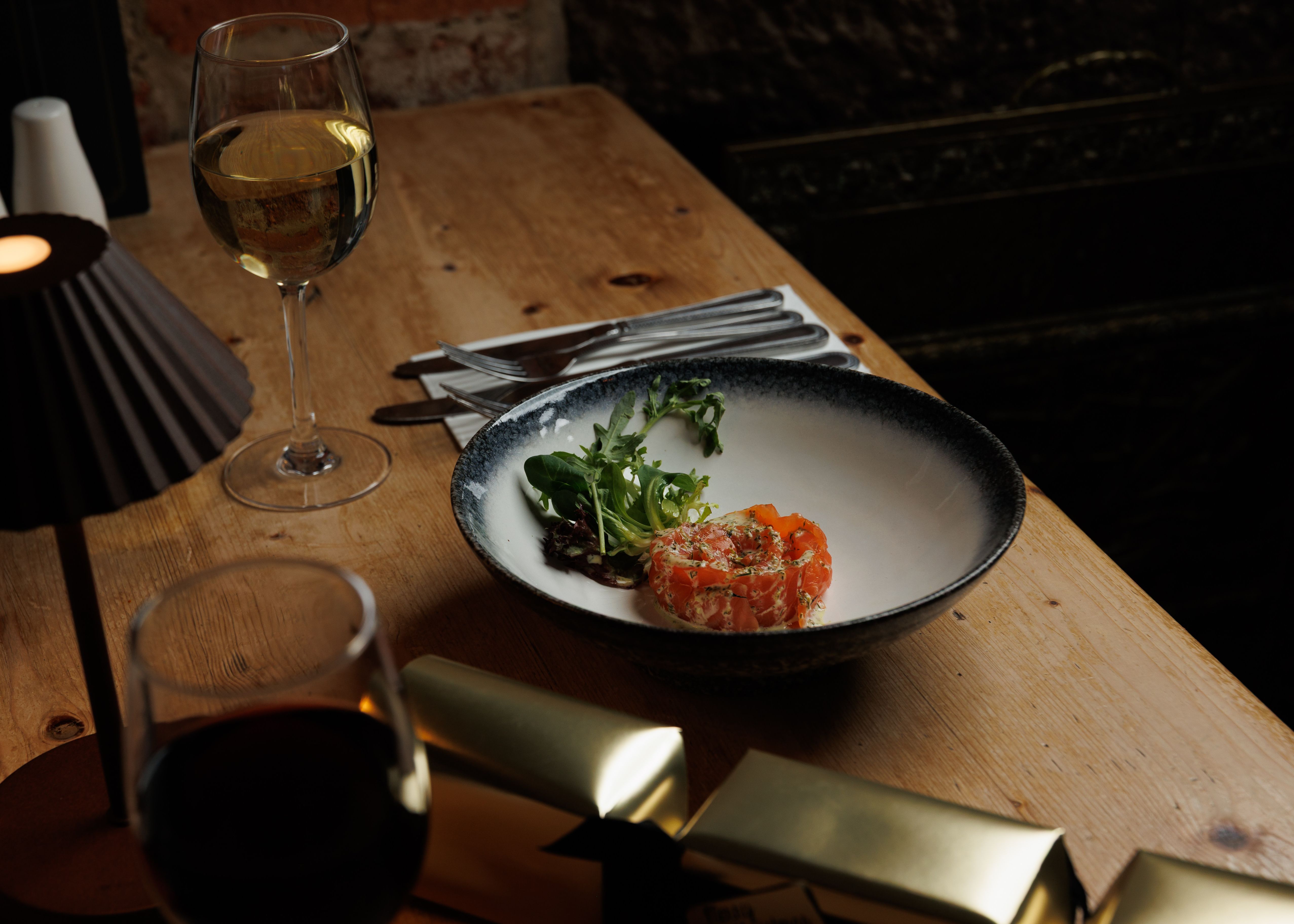 A plate with smoked salmon and salad greens on a wooden table, with wine glasses and cutlery nearby.