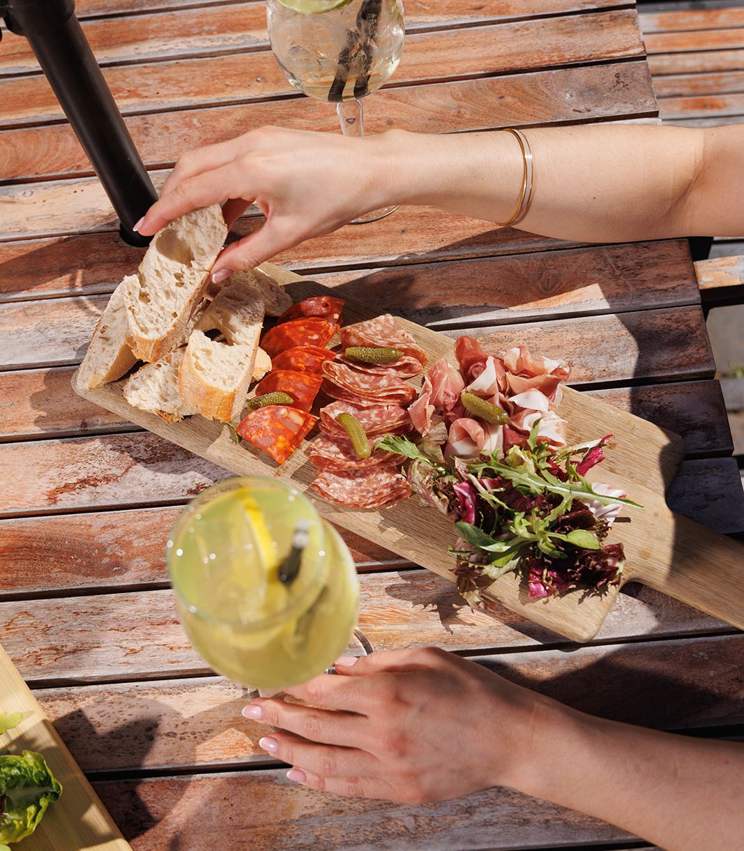 Hand reaching for bread on a charcuterie board with assorted meats and greens, with drinks on a wooden outdoor table.