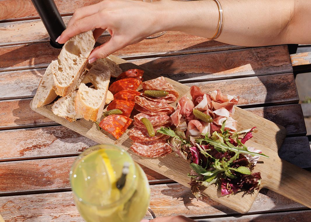 Hand reaching for bread on a charcuterie board with assorted meats and greens, with drinks on a wooden outdoor table.