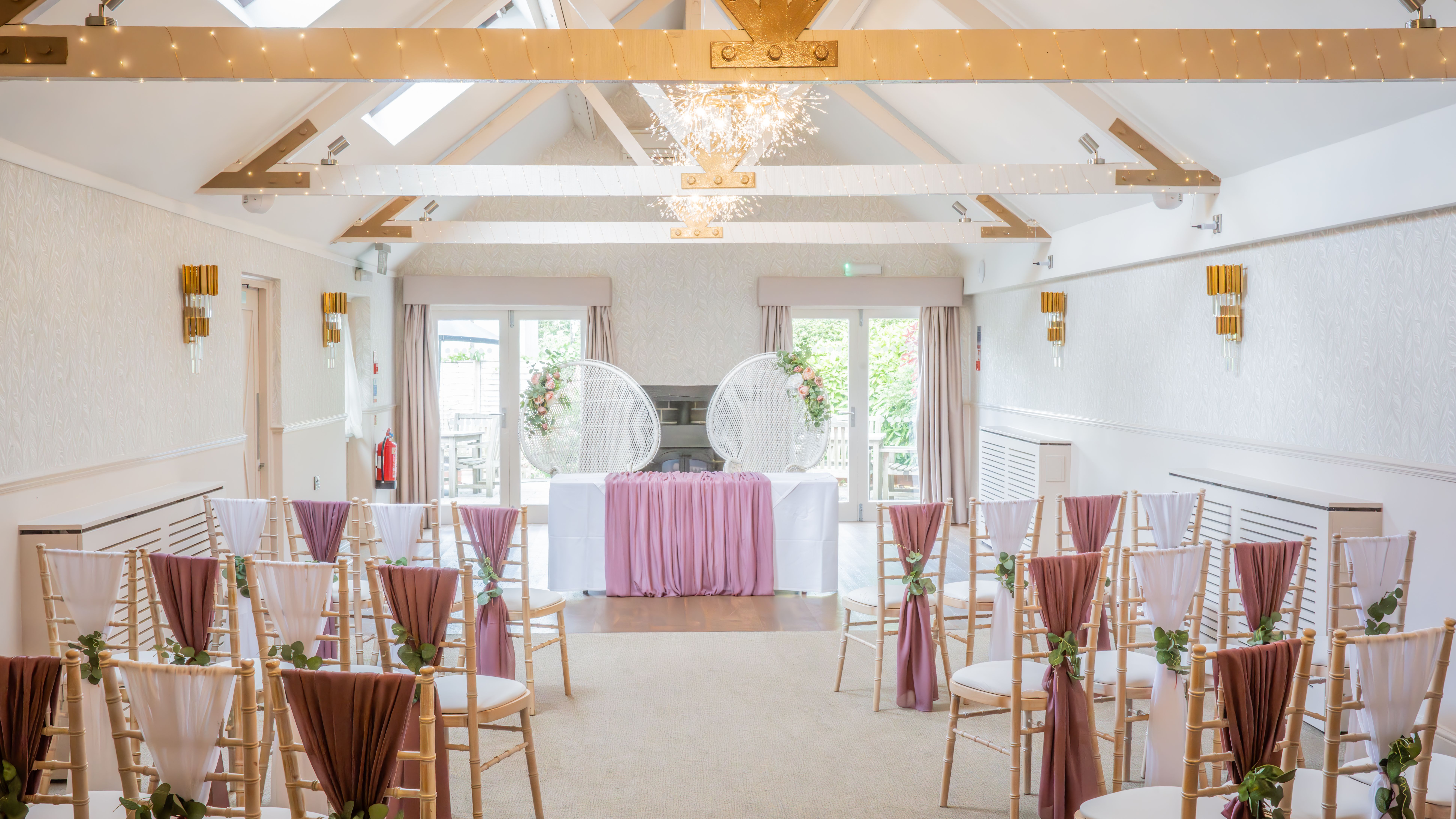 Beautiful indoor wedding ceremony setup with chairs draped in pink and white fabric, floral arrangements, and decorative lighting.