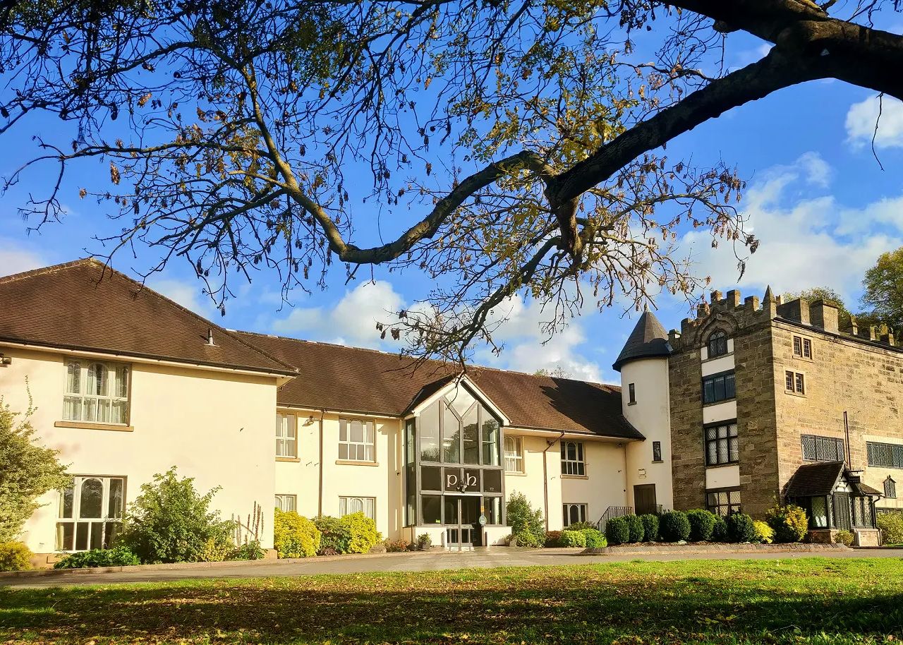 Large building with cream-colored walls and a stone tower under a blue sky, framed by tree branches.