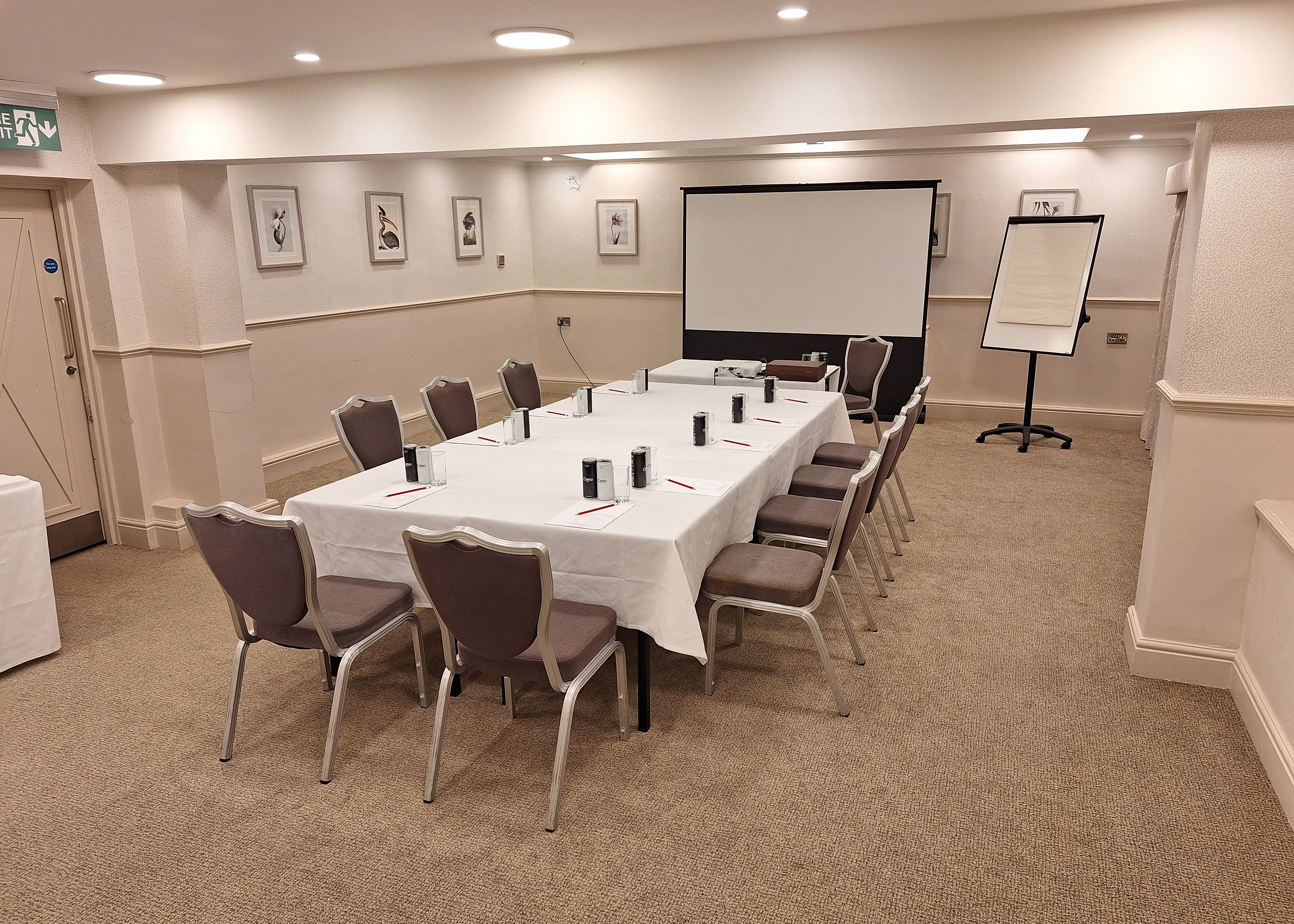 Conference room setup with a rectangular table, chairs, projector screen, and flipchart