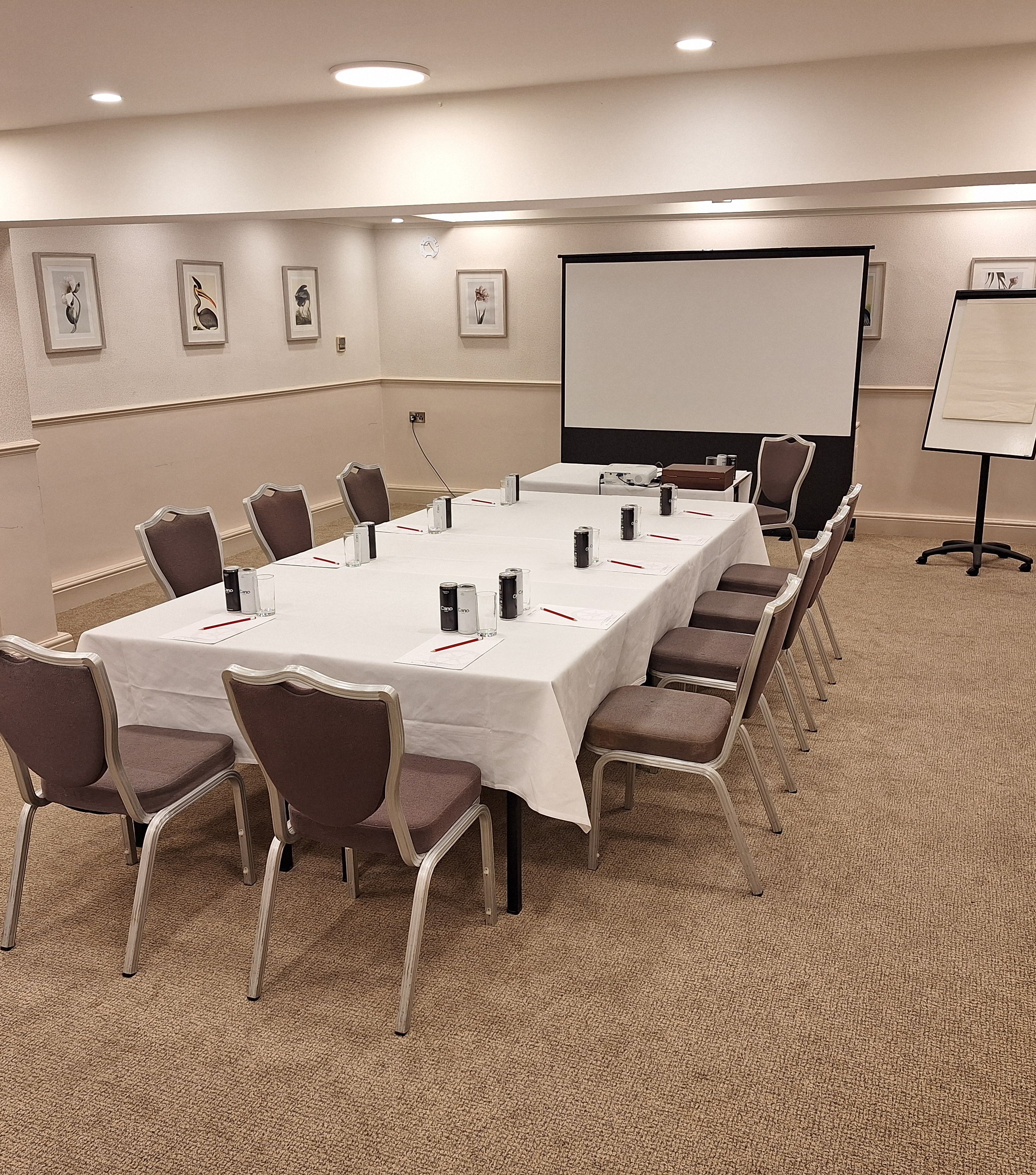 Conference room setup with a rectangular table, chairs, projector screen, and flipchart