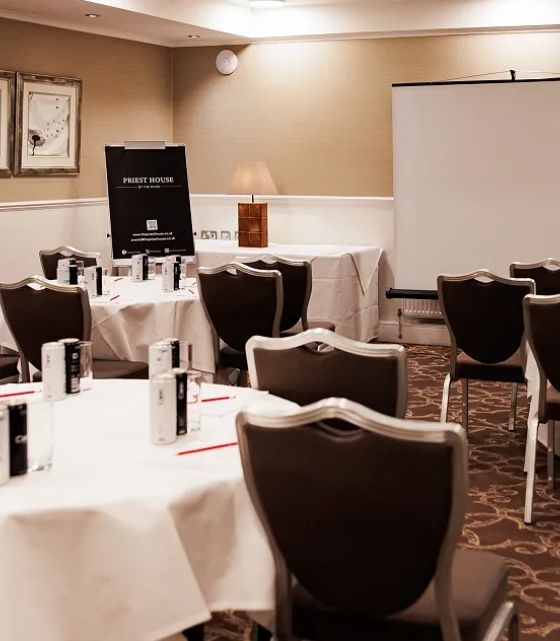 Conference room with round tables, chairs, a projector screen, and writing materials set up for a meeting.
