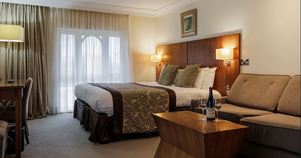 Modern hotel room with a large bed, sofa, wooden coffee table, and natural light coming through sheer curtains.