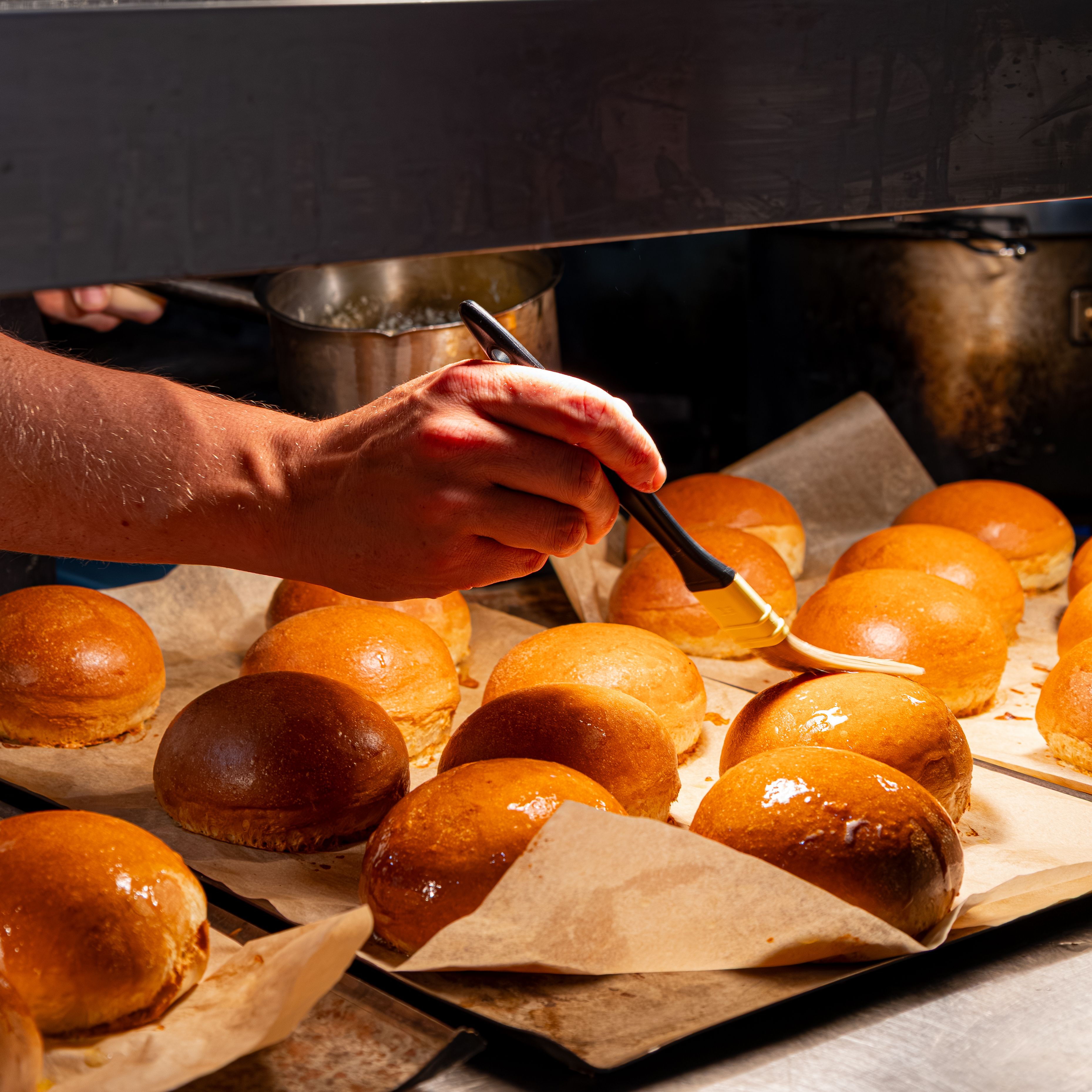 A person brushing glaze onto freshly baked bread rolls in a kitchen.