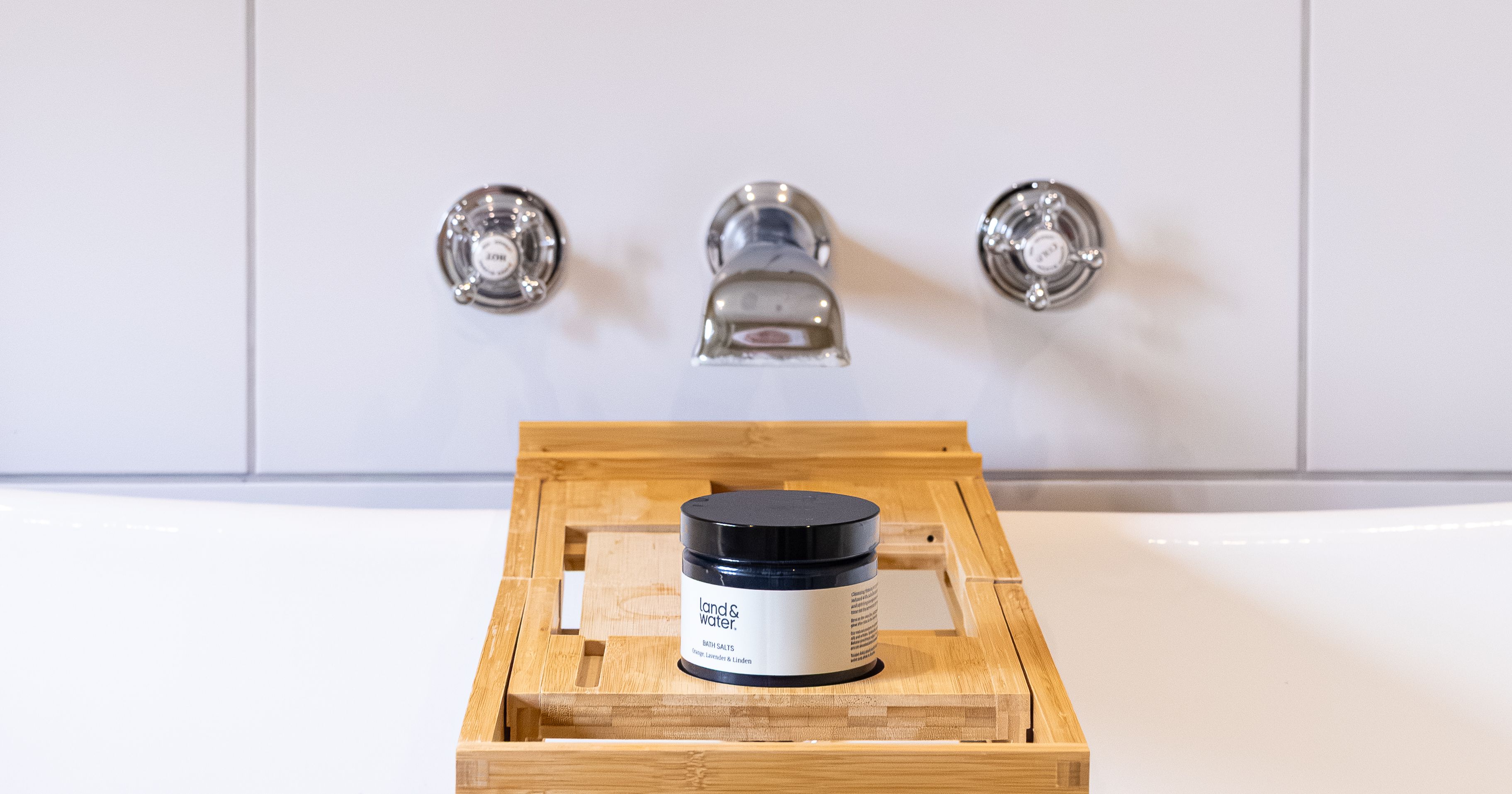 A jar of Land & Water product on a wooden bath tray over a white bathtub.