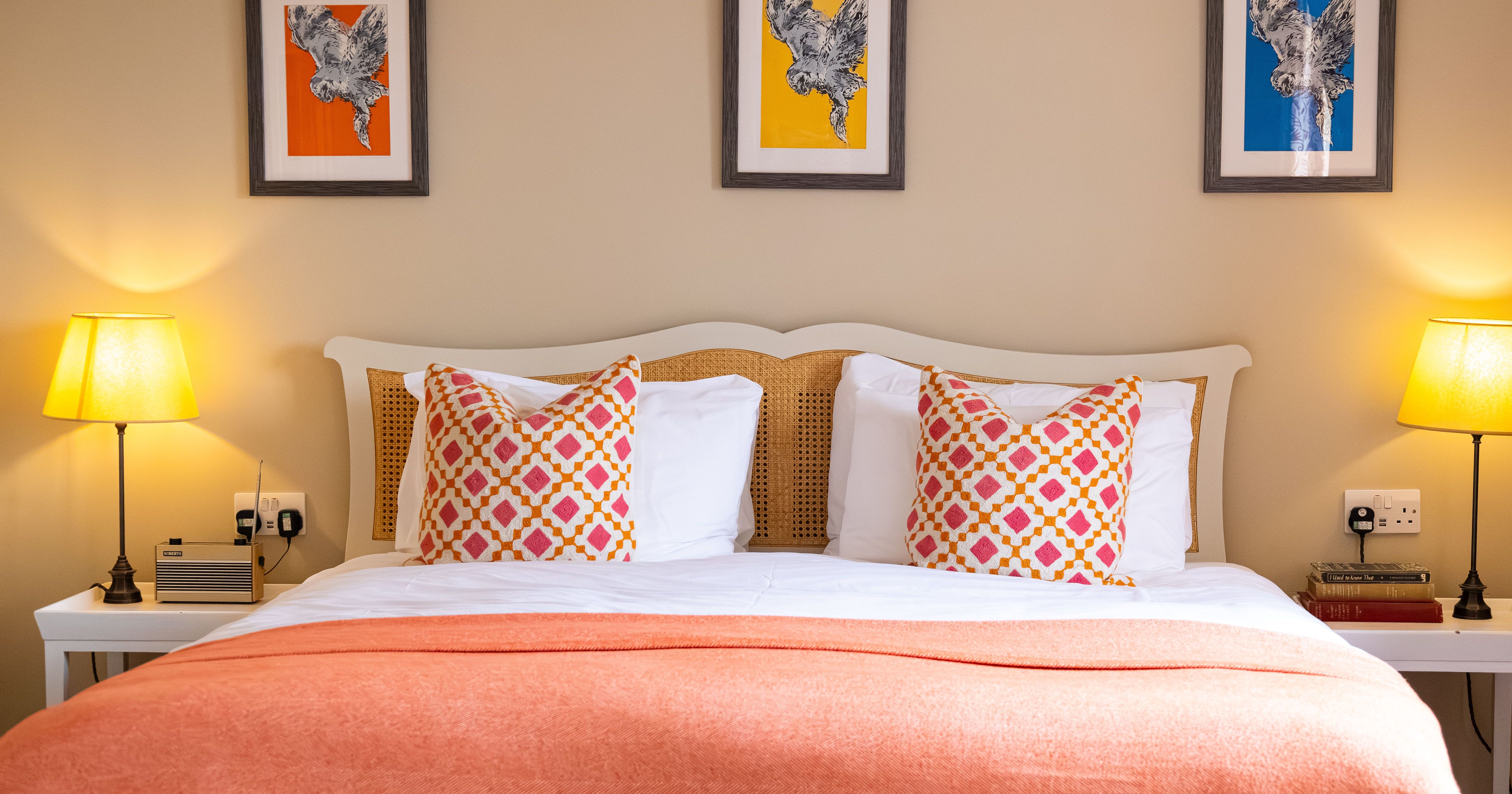 A cozy bed with colorful pillows and orange blanket, framed by two bedside tables with lamps and three animal art prints on the wall above.