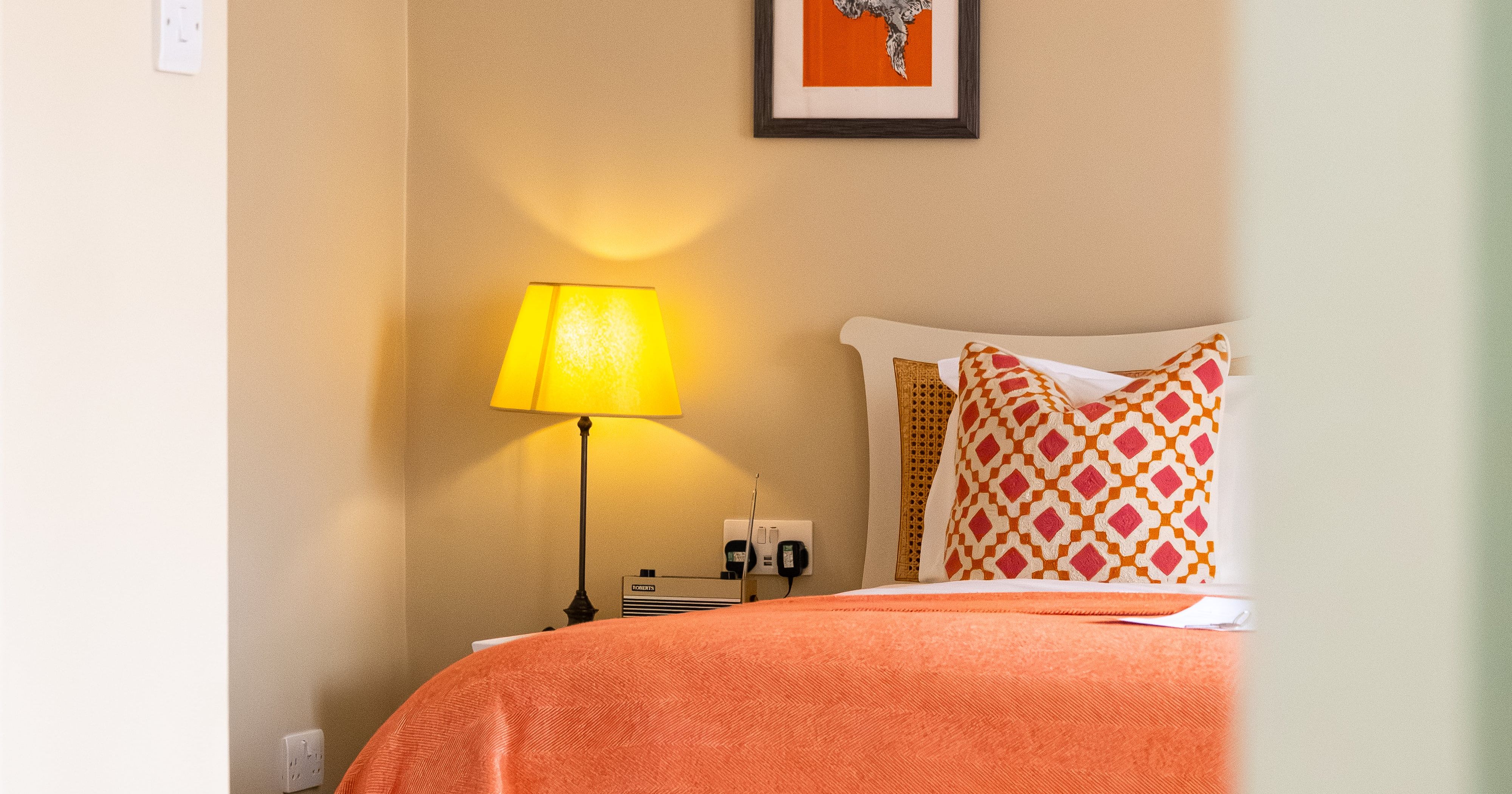 Cozy bedroom with orange bedspread, patterned pillow, lamp, and framed artwork on the wall.