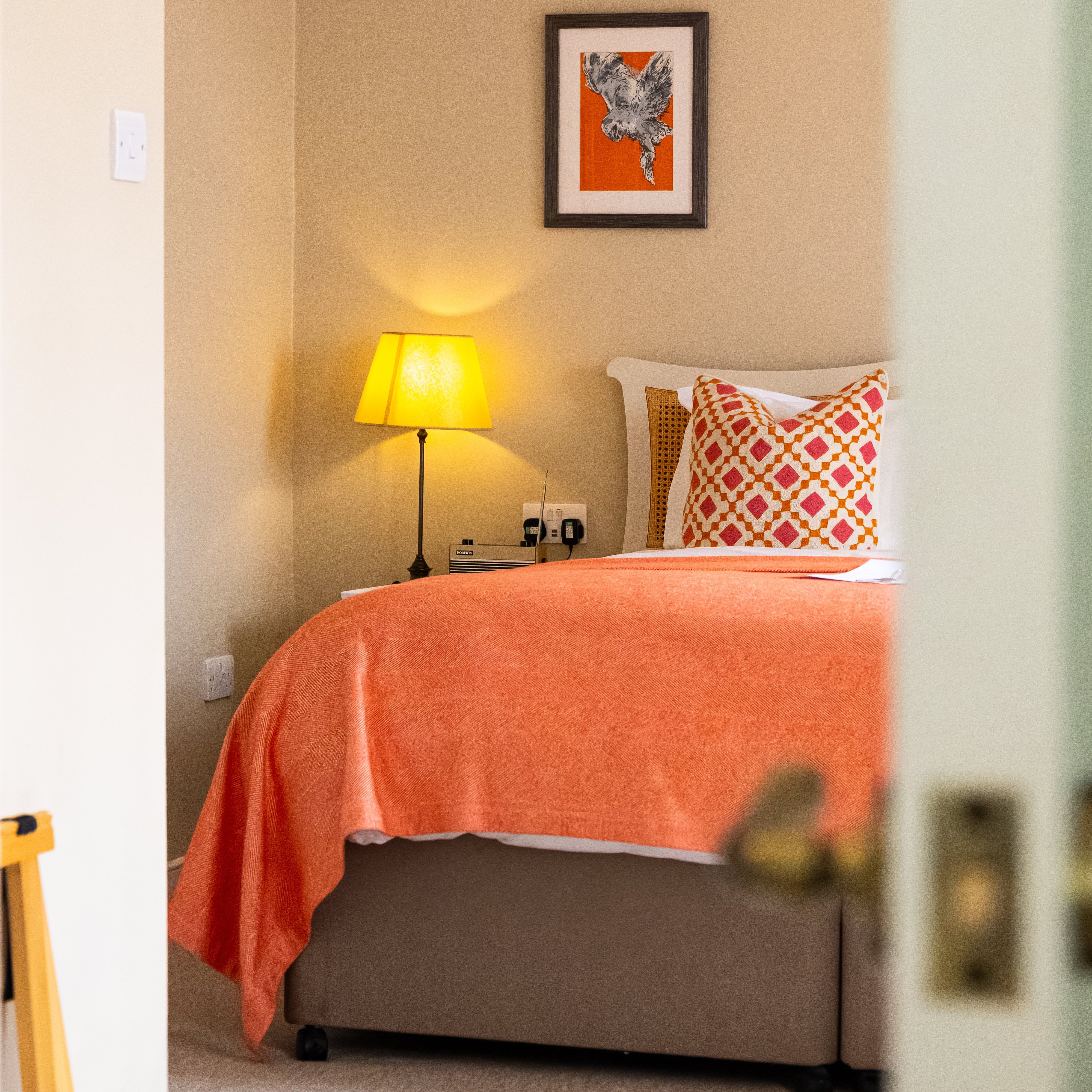 Cozy bedroom with orange bedspread, patterned pillow, lamp, and framed artwork on the wall.
