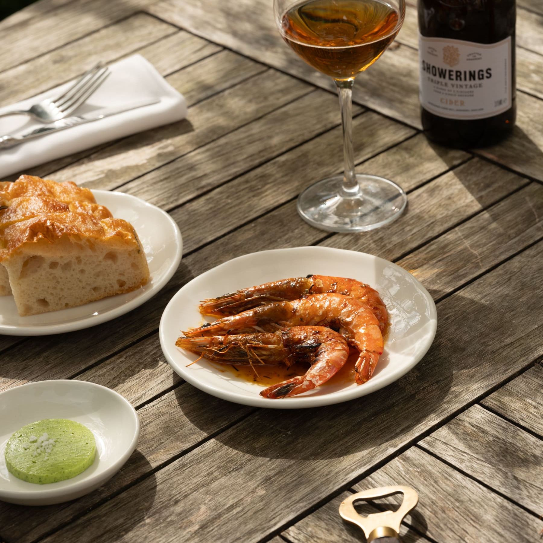 A wooden table set with grilled shrimp on a plate, a piece of bread, a small dish with a green spread, a glass of cider, and a bottle of Showerings Triple Vintage cider.