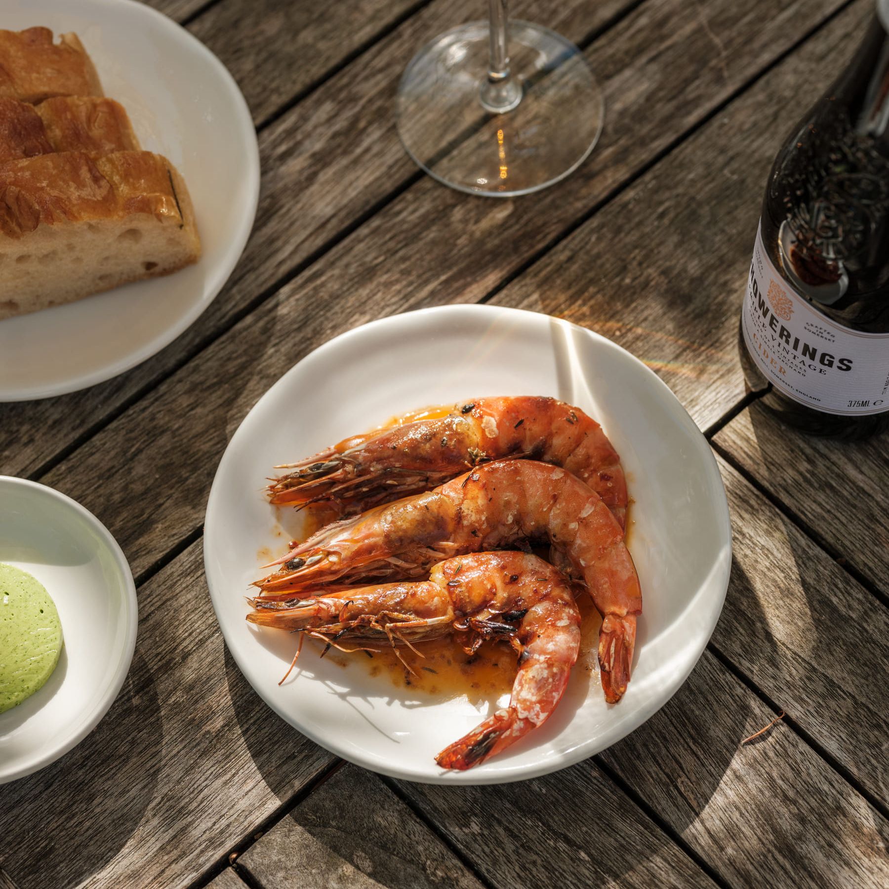 A plate of cooked prawns on a wooden table with a glass of wine, bread, and a green macaron.