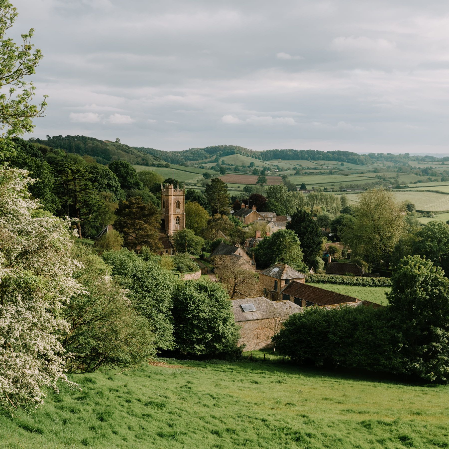 A picturesque English countryside village with a church tower, surrounded by green fields, trees, and rolling hills.