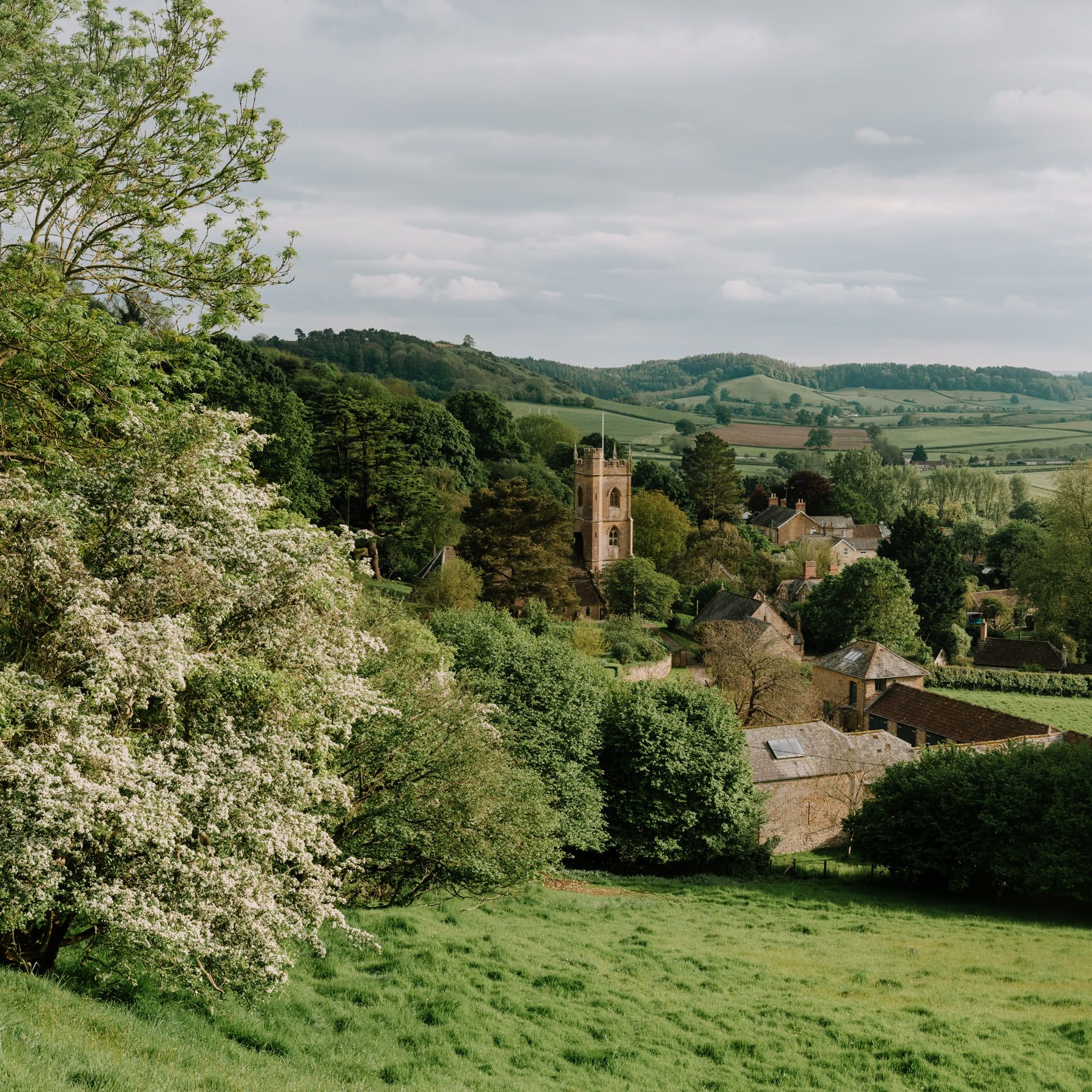 A picturesque English countryside village with a church tower, surrounded by green fields, trees, and rolling hills.