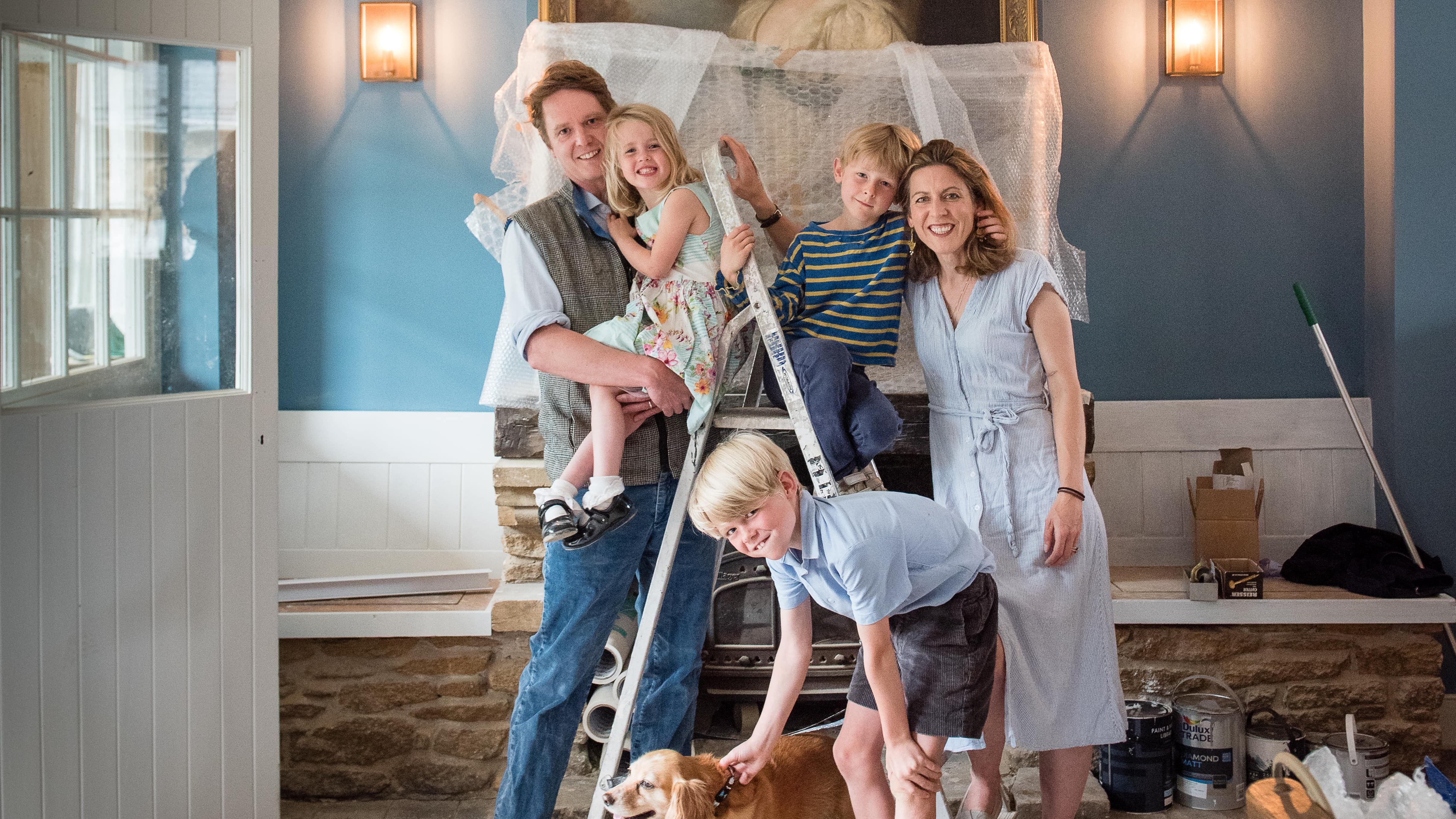 Family posing in a room under renovation with a dog and a ladder, with a large portrait painting on the wall behind them.