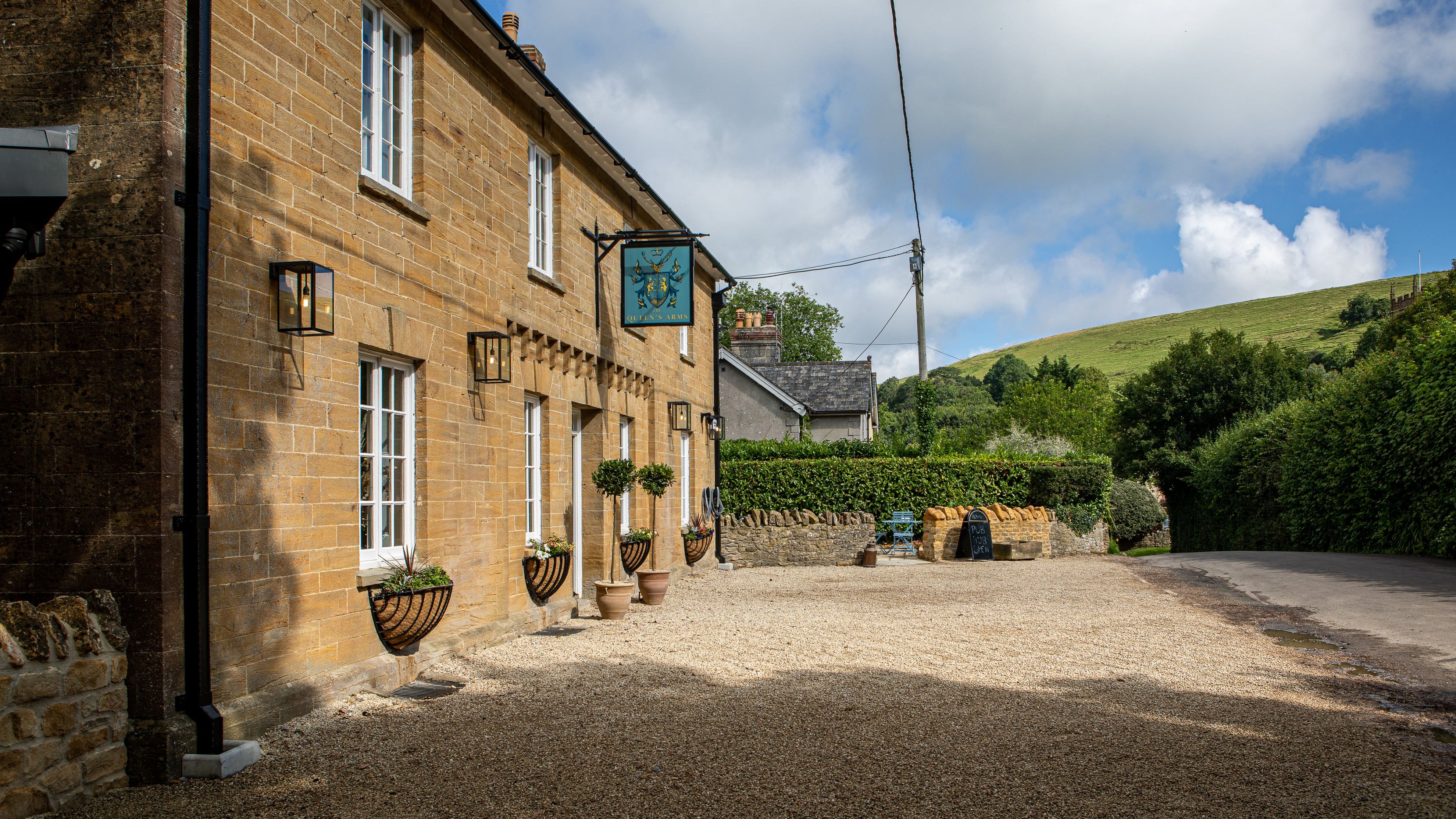 Traditional stone building with white windows and a pub sign, set along a rural road with green hills in the background.