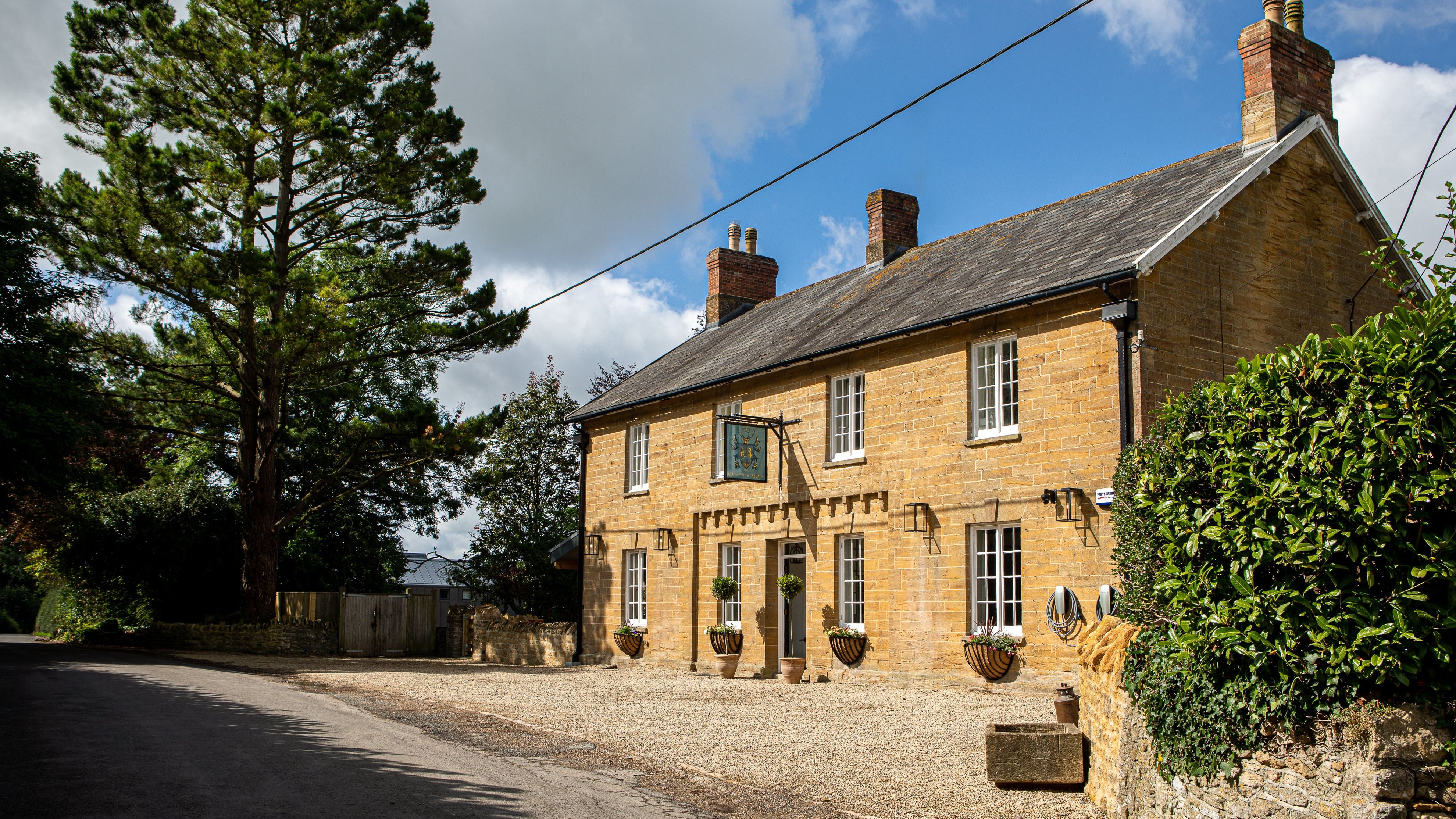 Traditional stone house with white-framed windows and a gravel driveway, surrounded by greenery and a large tree.