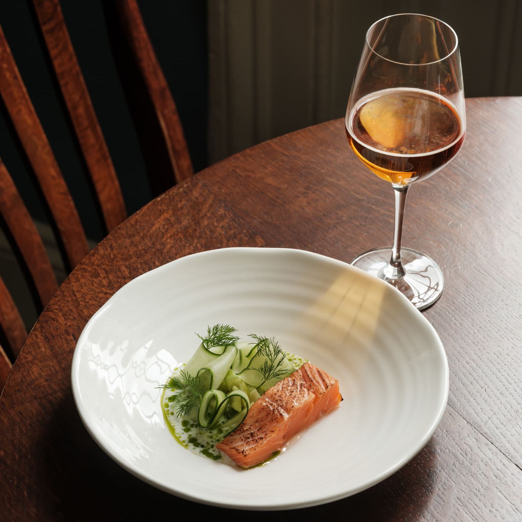 A plated piece of salmon with cucumber and herbs, served with a glass of amber-colored wine on a wooden table.