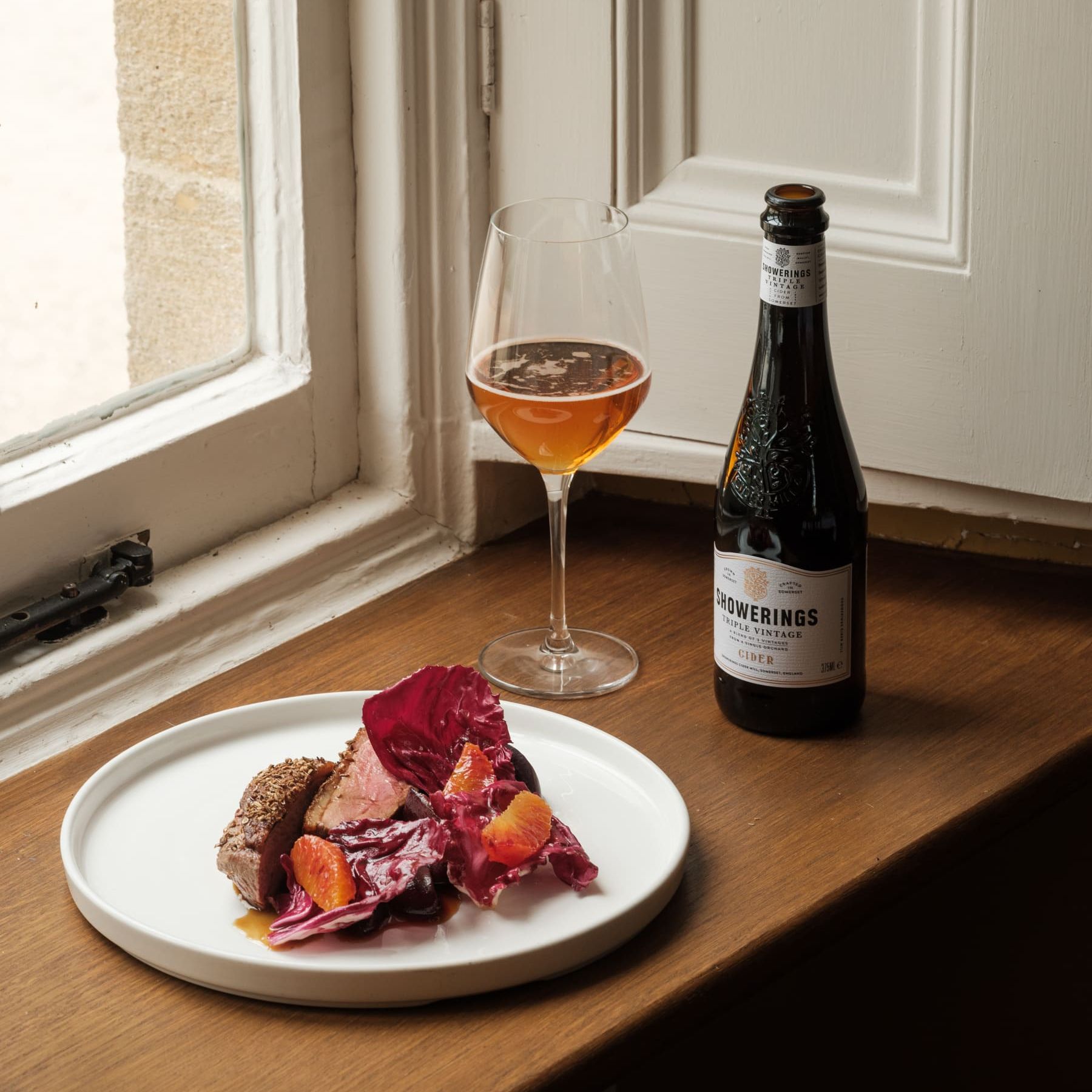 A plated gourmet dish with a glass of cider and a bottle of Showerings cider on a wooden windowsill.