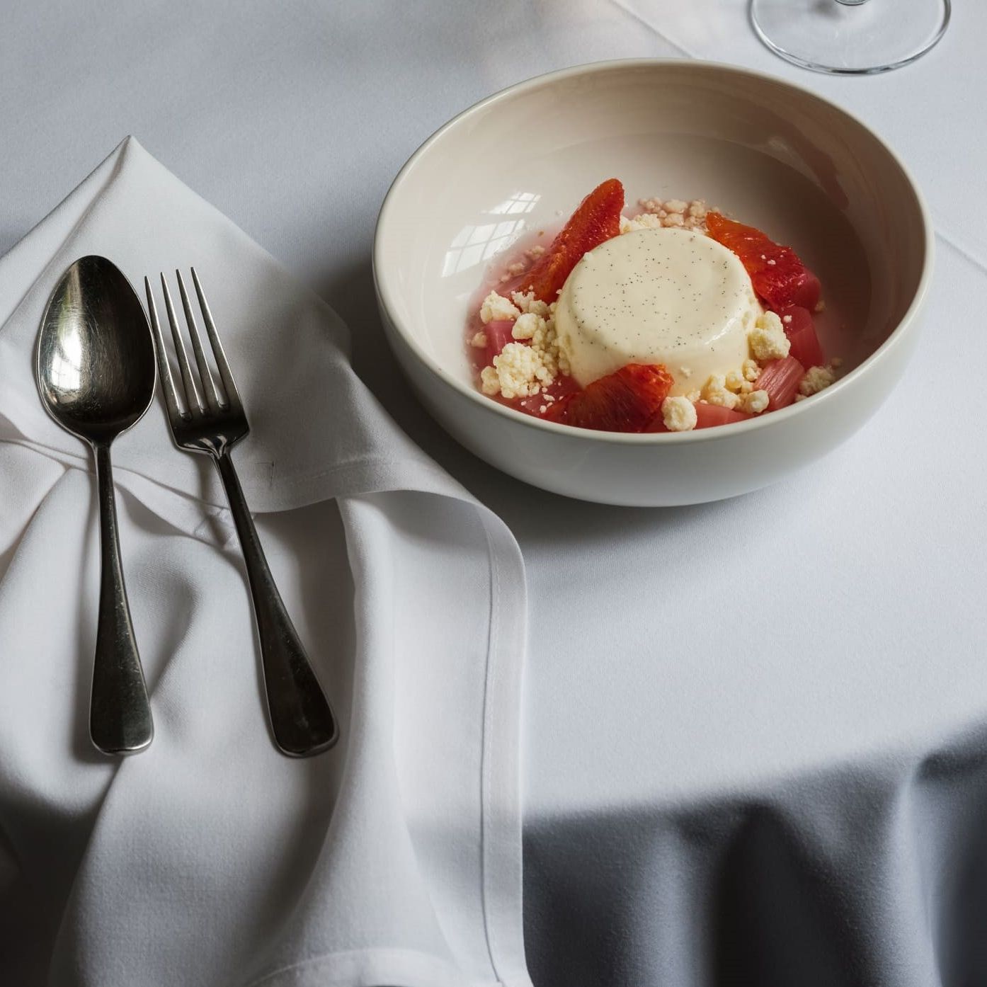 A bowl of panna cotta dessert garnished with fruit and crumbles, set on a white tablecloth beside a fork and spoon.