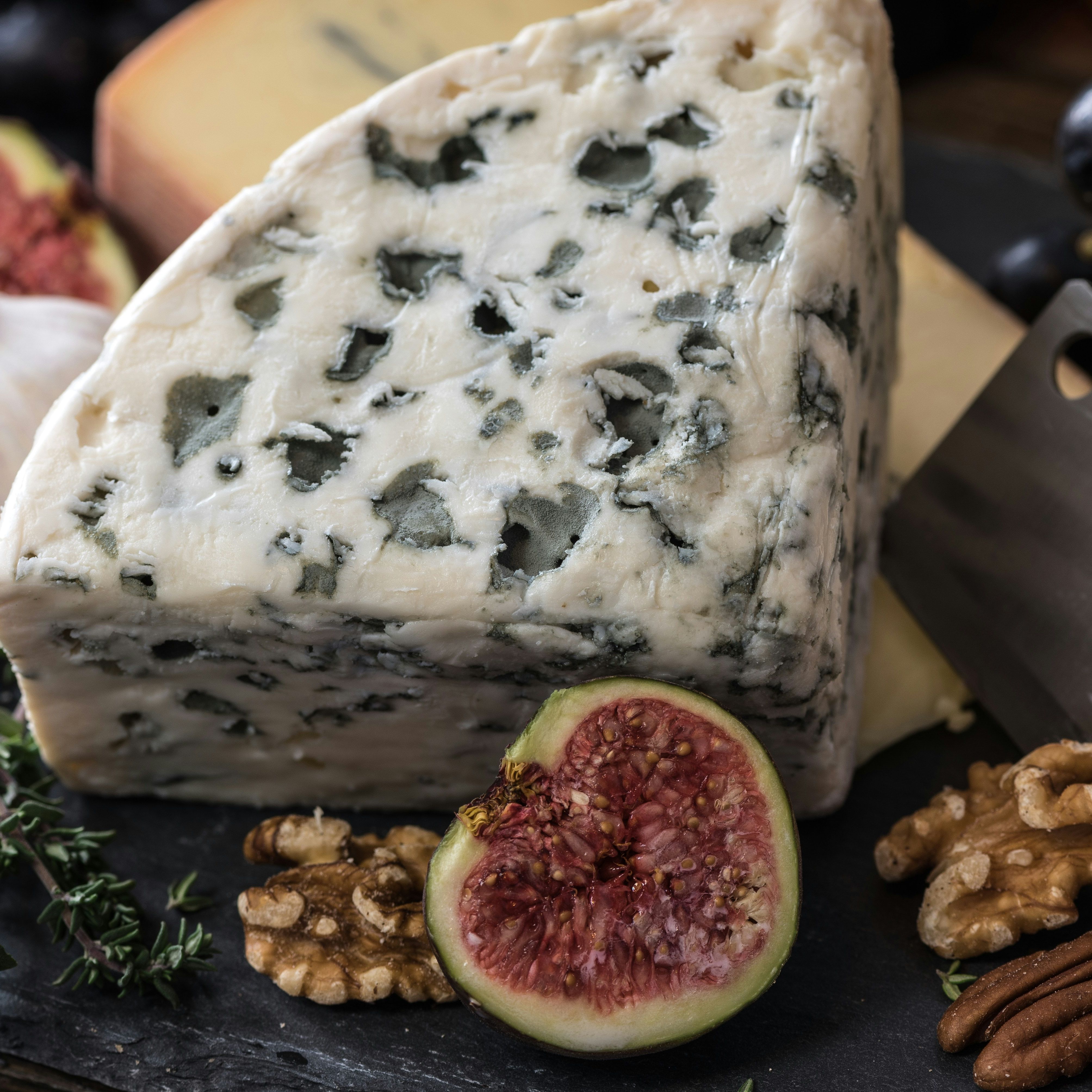 A wedge of blue cheese on a slate platter with figs, walnuts, garlic, herbs, and a cheese knife