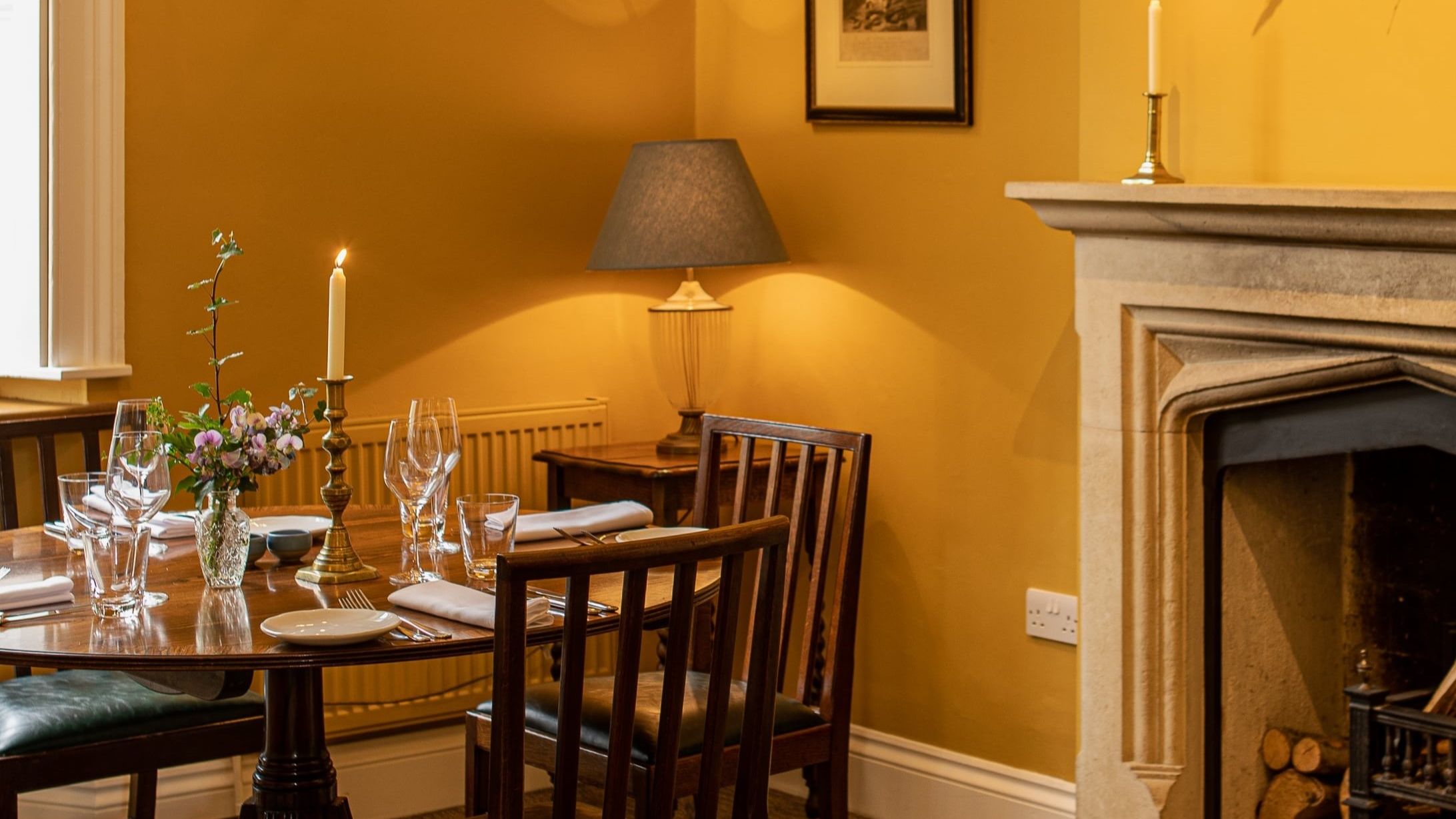 Cozy dining room with wooden table and chairs, set for a meal, near a lit fireplace and decorated with flowers and candles.