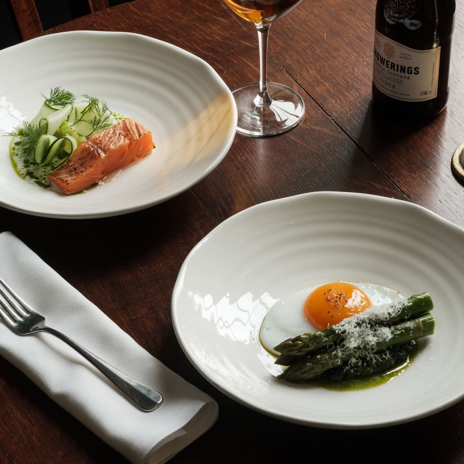 Two plates of gourmet food on a wooden table, one with salmon and cucumber, the other with asparagus and a sunny side up egg.