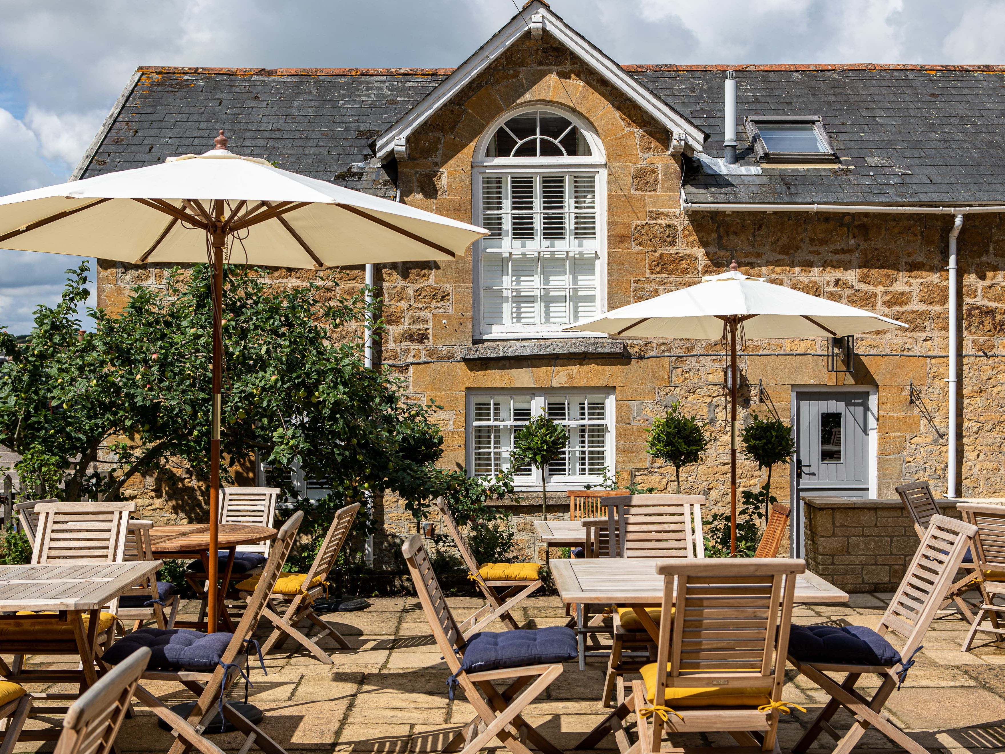 Outdoor patio area with wooden tables and chairs beneath umbrellas in front of a stone house.
