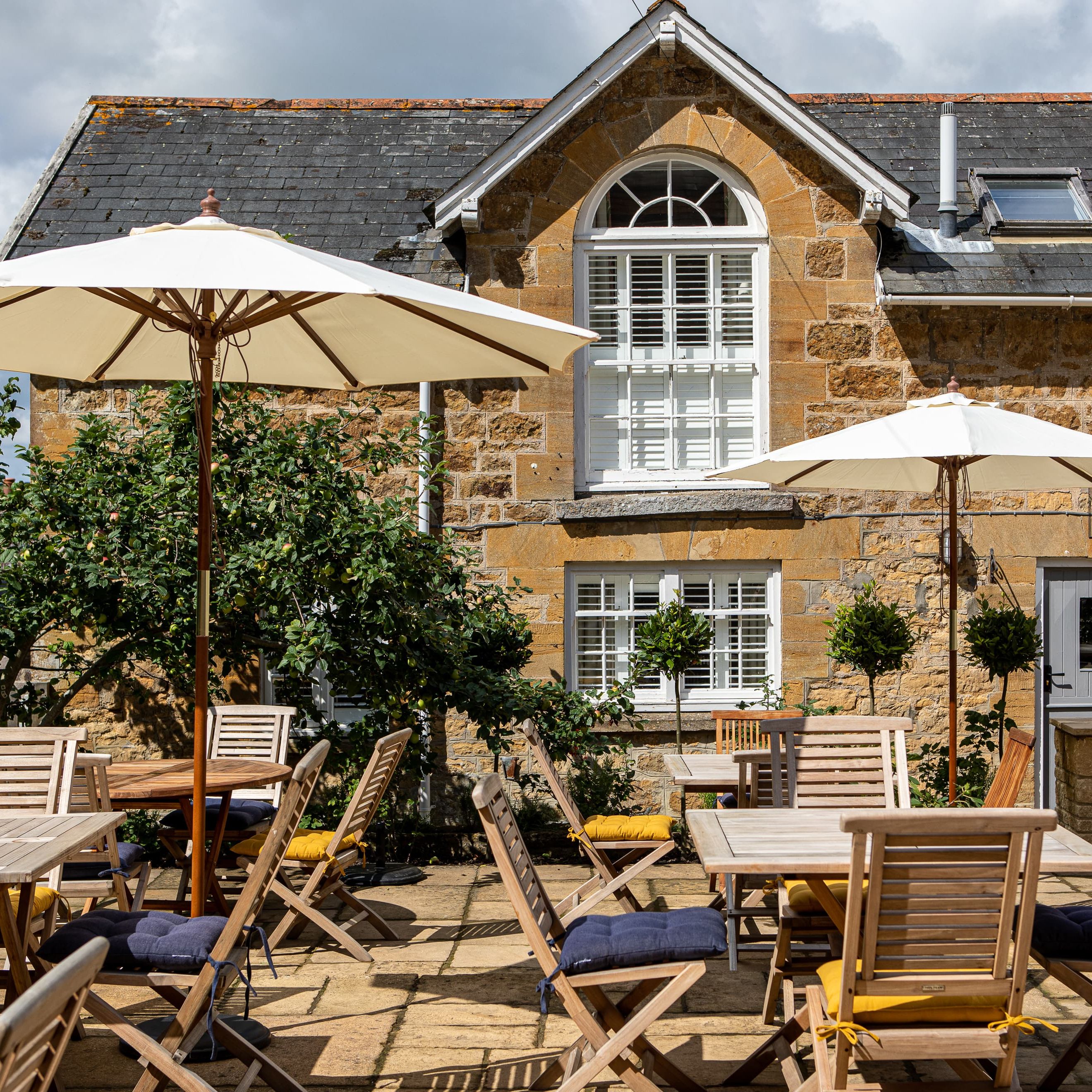 Outdoor patio area with wooden tables and chairs beneath umbrellas in front of a stone house.