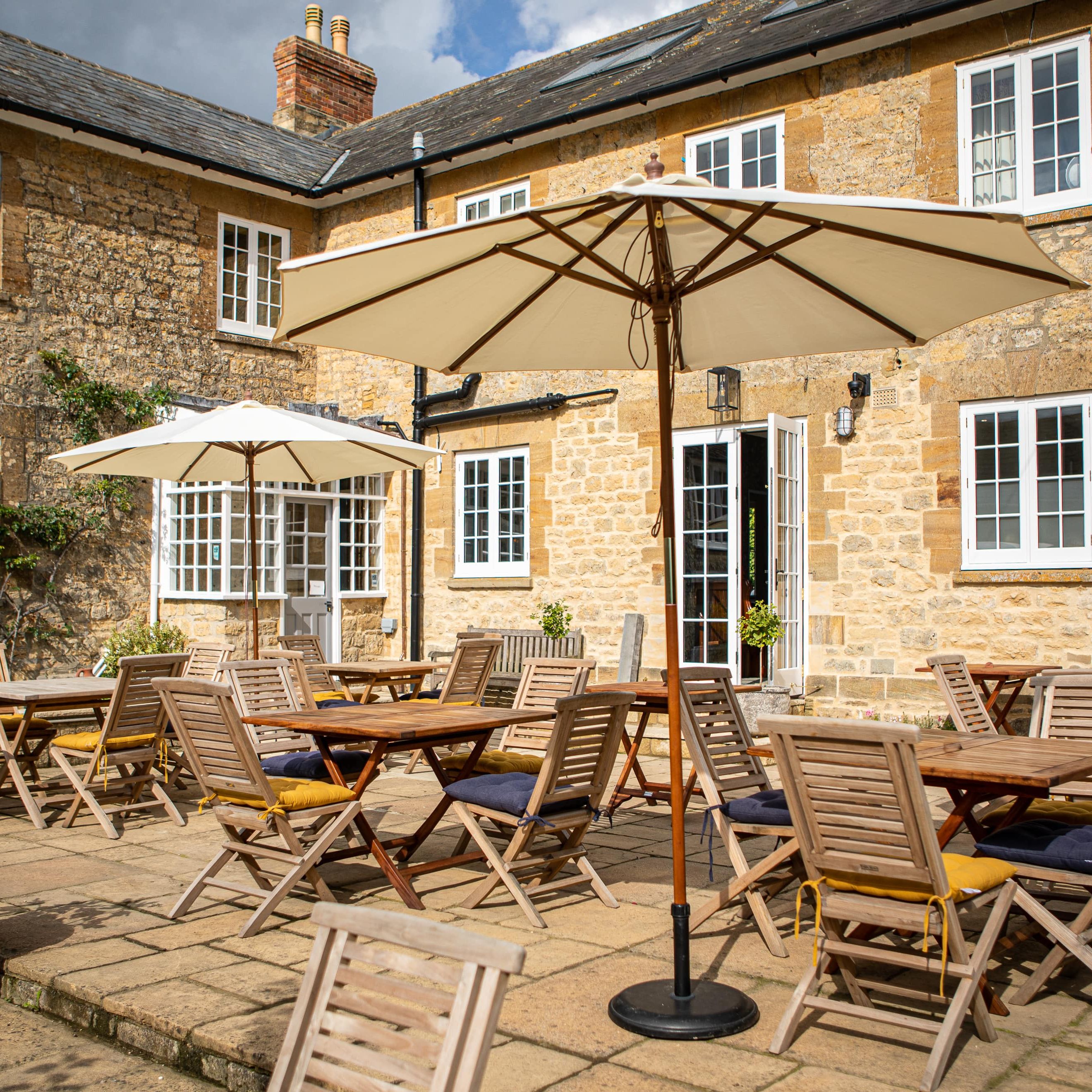 Outdoor patio seating area with wooden tables and chairs under large umbrellas in front of a stone building.
